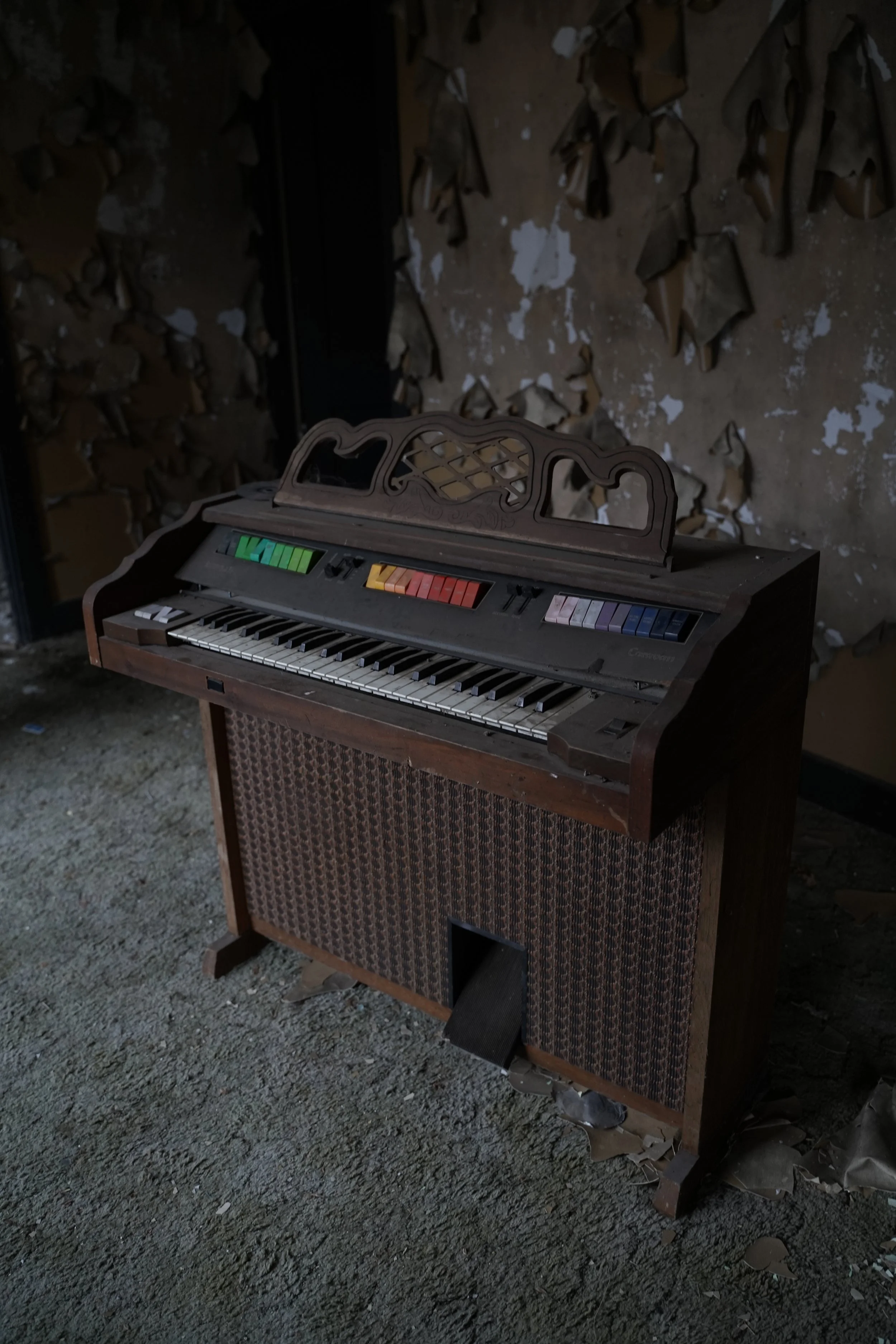 An antique electric organ with colorful keys and a decorative wooden railing on top, placed against a wall with peeling paint and various hanging leather pieces.