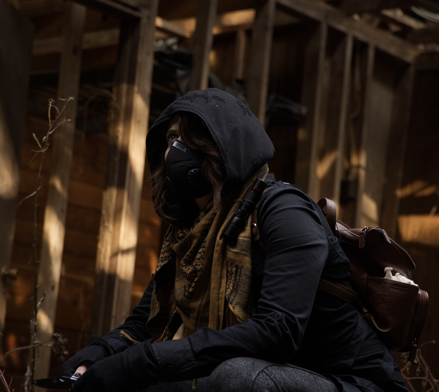A woman wearing a black hoodie, face mask, and scarf, crouching indoors with a wooden structure in the background, carrying a backpack.