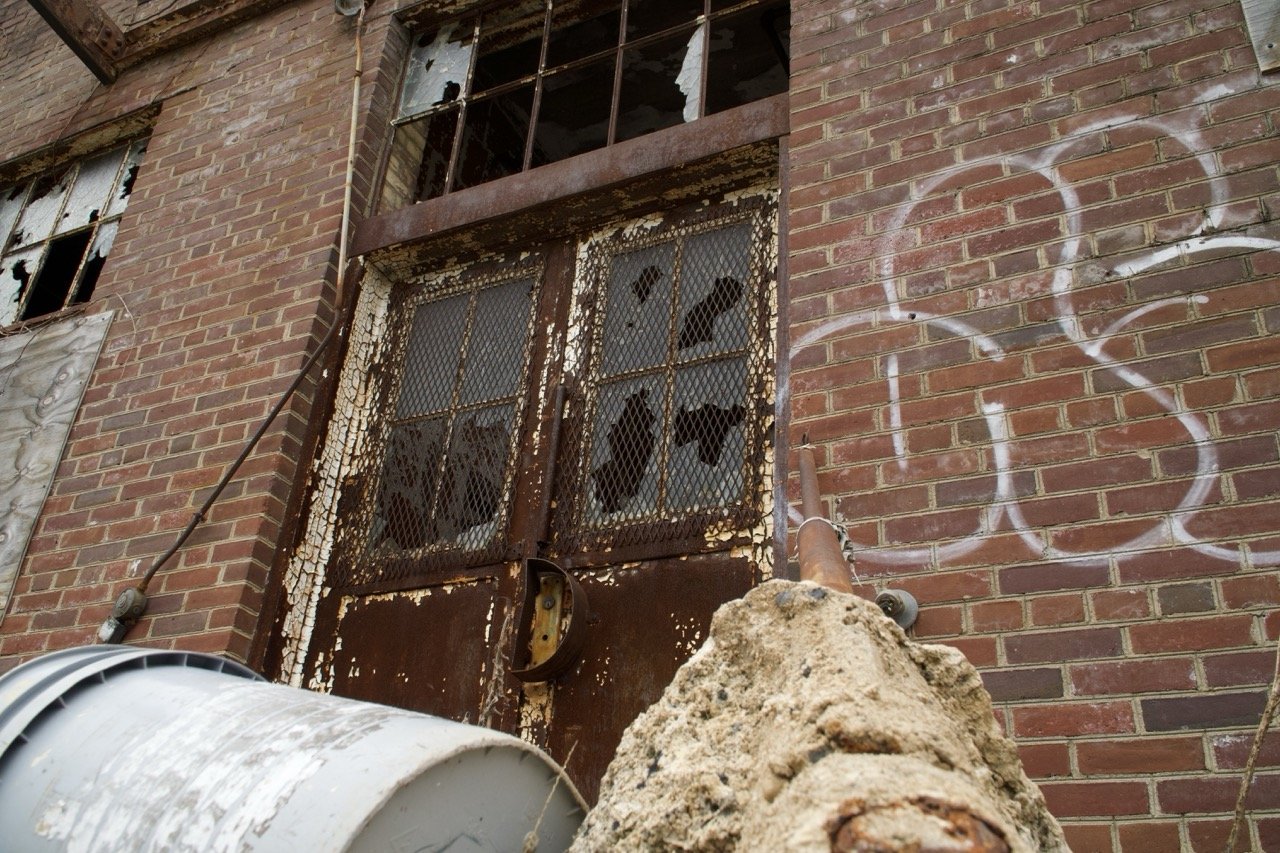 An old, rusty fire escape with chain-link fencing on a brick building, broken windows, graffiti, and a large pipe or drain in the foreground.