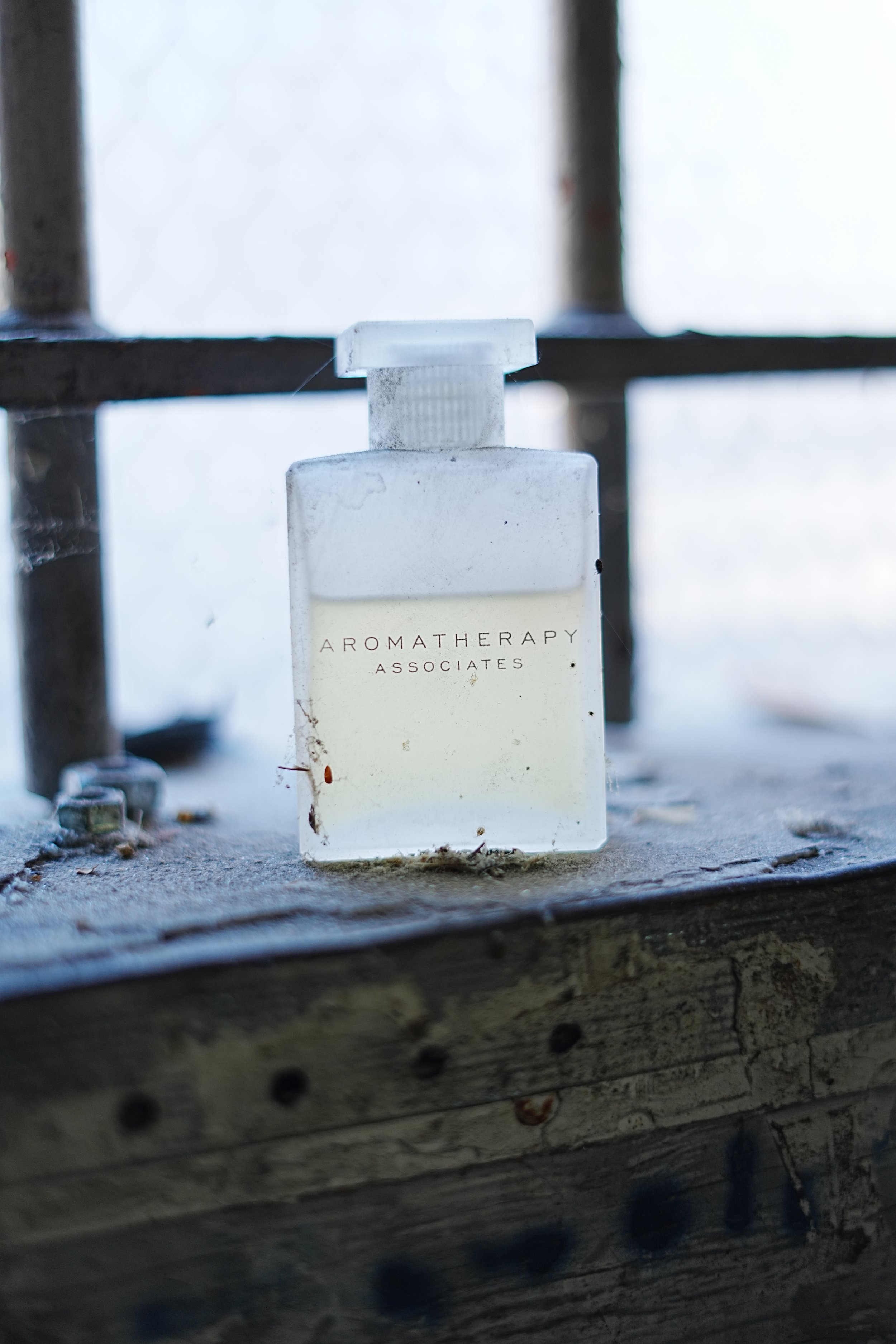 Close-up of a white aromatherapy diffuser bottle labeled 'Aromatherapy Associates' sitting on a wooden windowsill with a spider web nearby, illuminated by natural light.