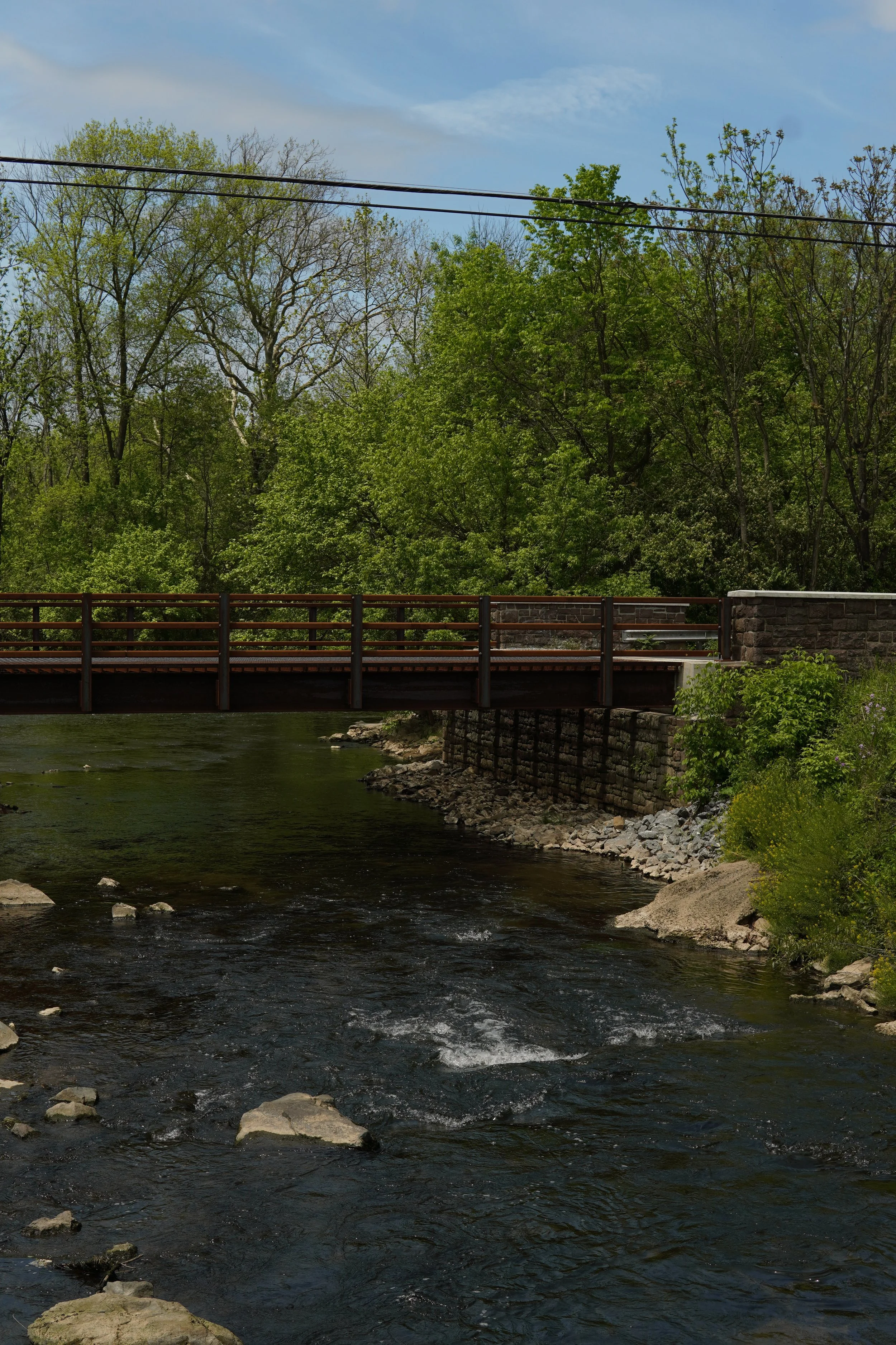 A scene of a river flowing beneath a wooden pedestrian bridge, with green trees on both sides and a blue sky with some clouds.