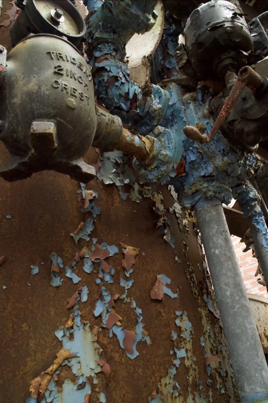 An old, rusty industrial valve with peeling blue paint and attached pipes, on a corroded metal surface.