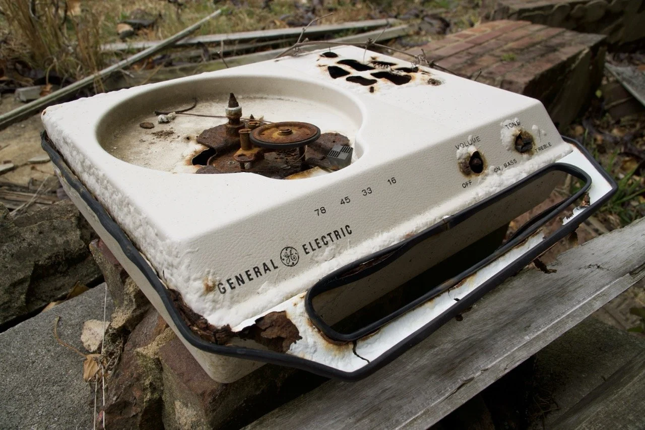 An old, rusty, and damaged electric stove top on a weathered wooden surface outdoors, with rust, black burn marks, and missing parts.