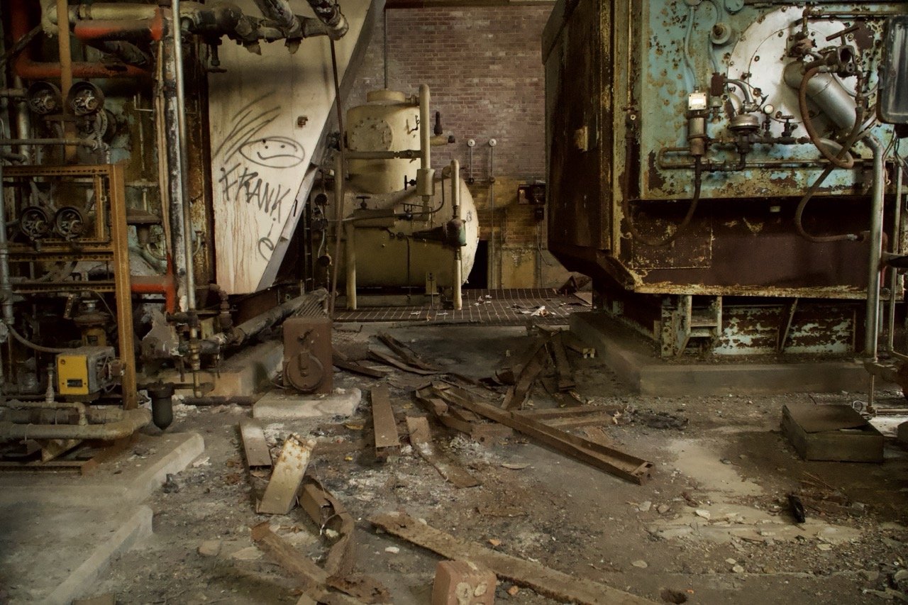 Abandoned industrial machinery and equipment in a gritty, rusty factory or workshop with scattered debris and graffiti on the wall.