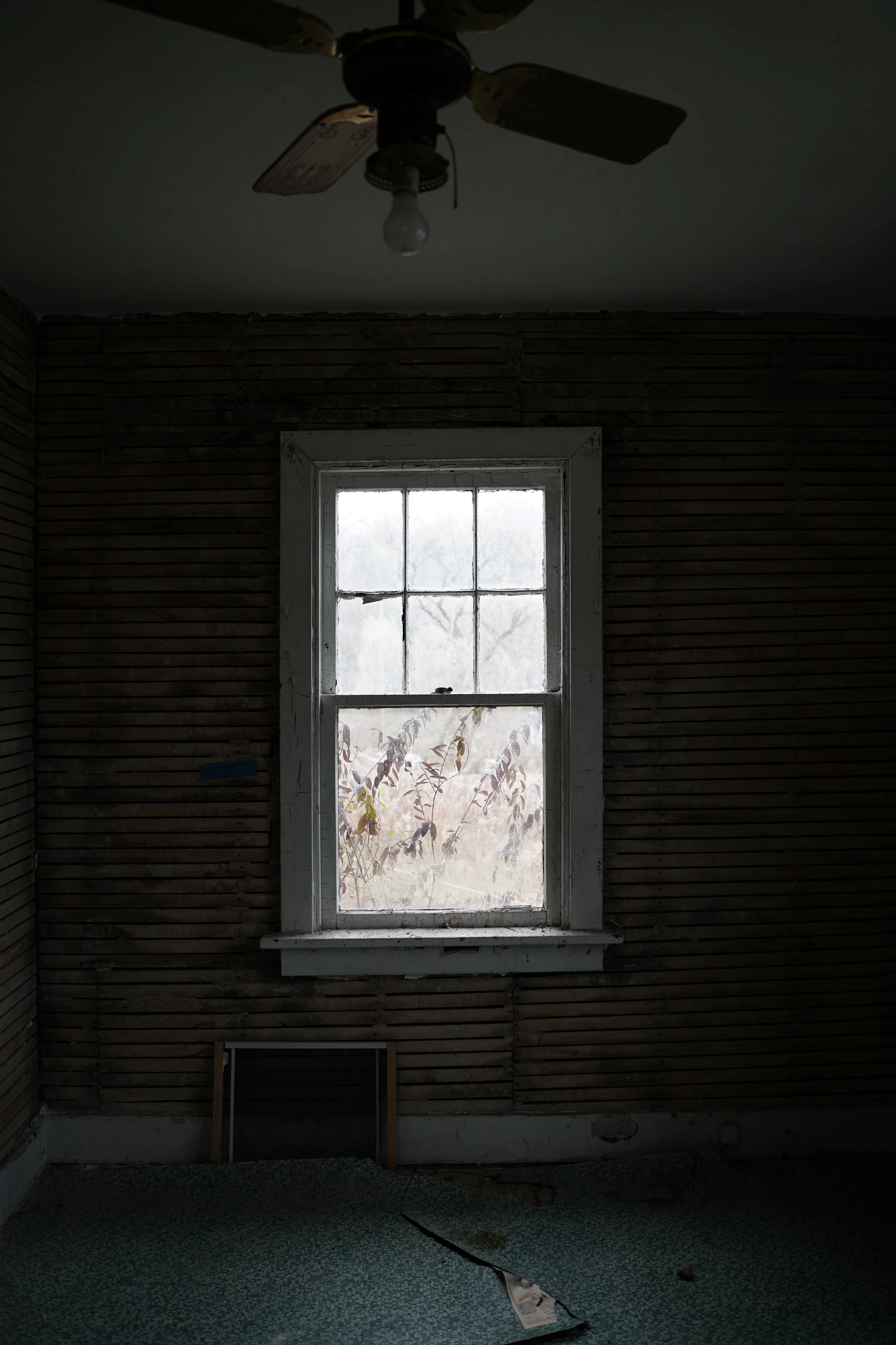 Dark, empty room with a window letting in light. The ceiling fan is visible with a light bulb. The wall is unfinished with wooden slats, and the floor has peeling wallpaper with a large crack and debris.