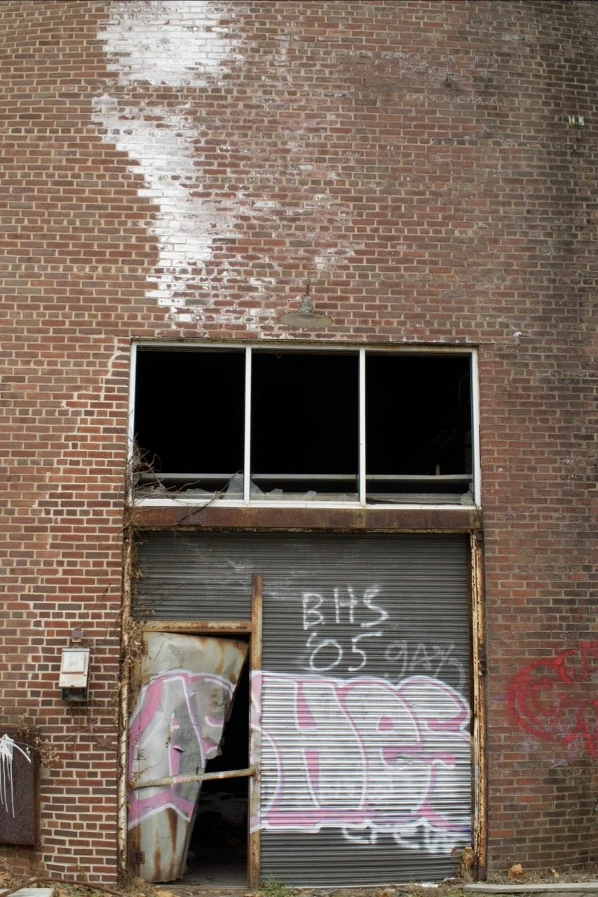 An old, abandoned brick building with graffiti on the roll-up garage door. The door is rusty and partially open, revealing a dark interior. There is graffiti on the door and above it, with the writing 'BHS 05 09 97' and some pink and white spray-pain