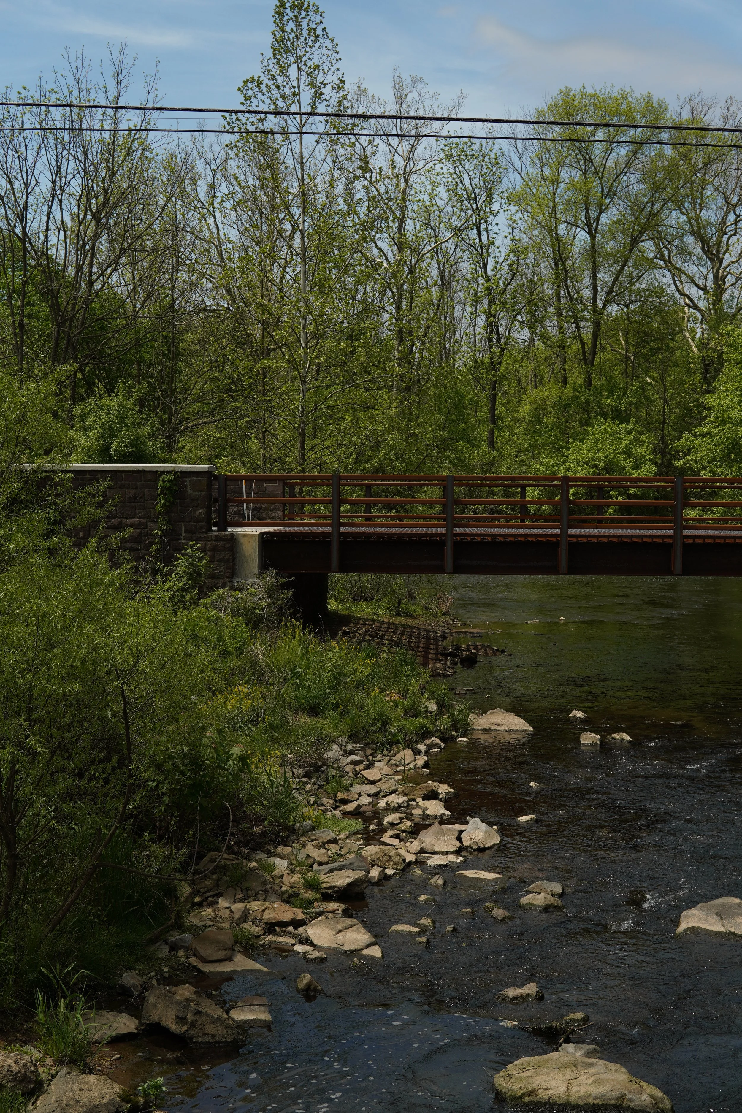 A river flowing under a bridge with trees in the background and power lines overhead.
