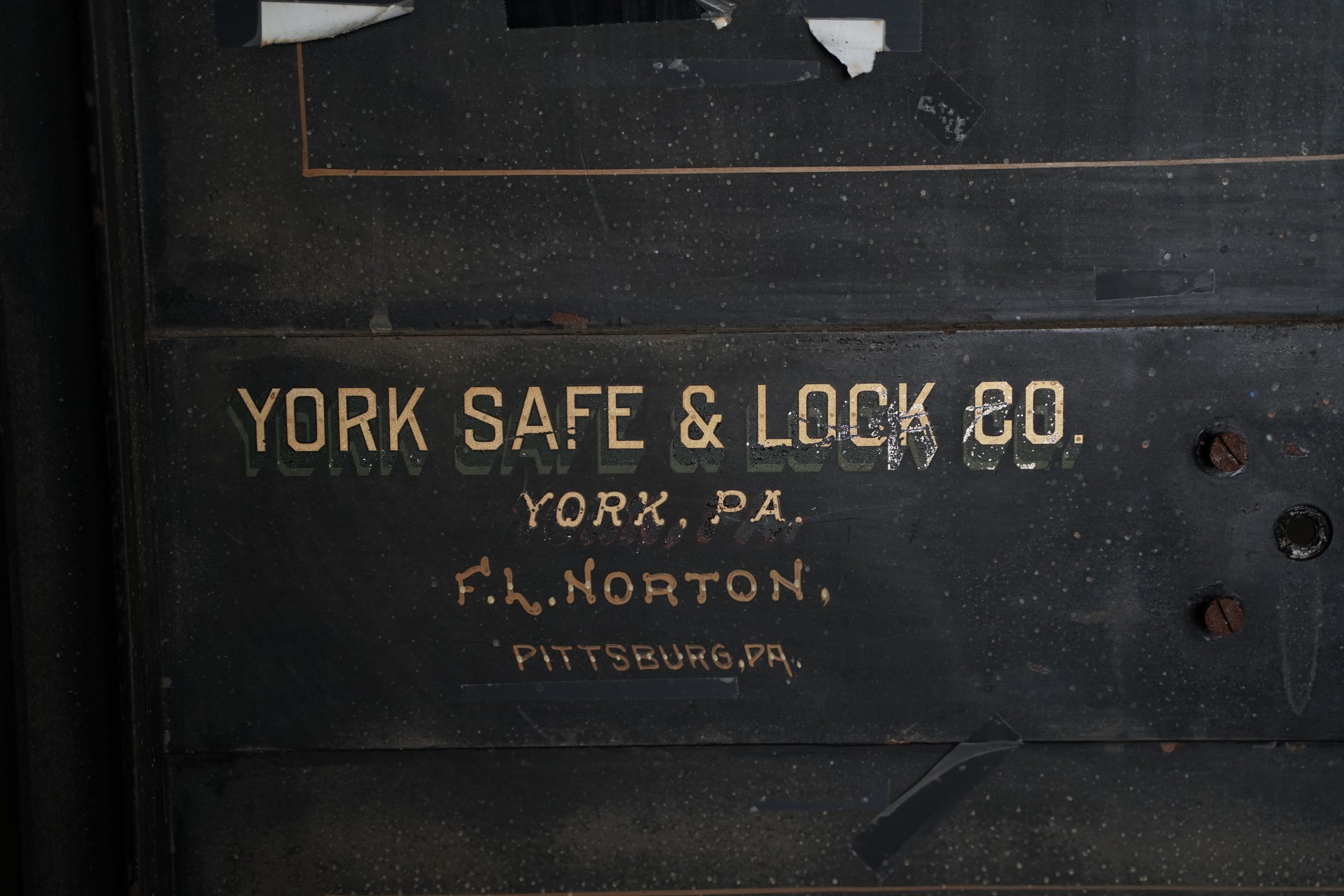 Close-up photo of an old, weathered metal safe with faded text that reads 'YORK SAFE & LOCK CO., YORK, PA, F. H. NORTON, PITTSBURG, PA.' The safe's surface is black with signs of rust and chipping paint.