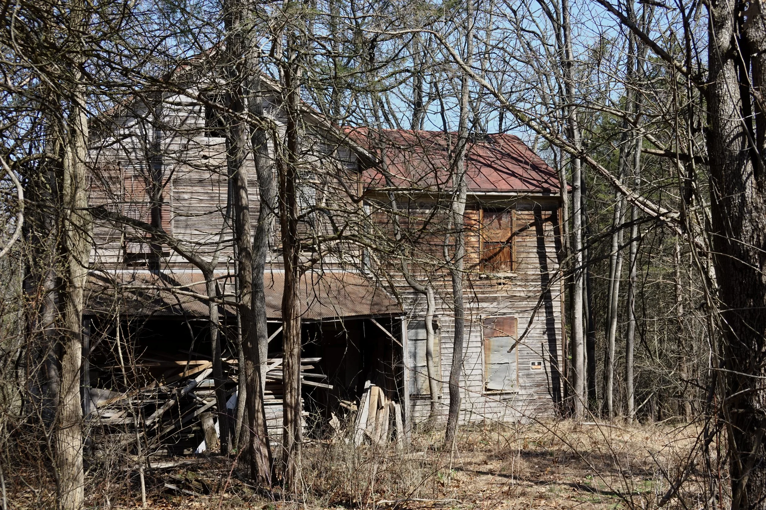 An abandoned wooden house surrounded by leafless trees in a wooded area, with a rusty roof and broken windows.