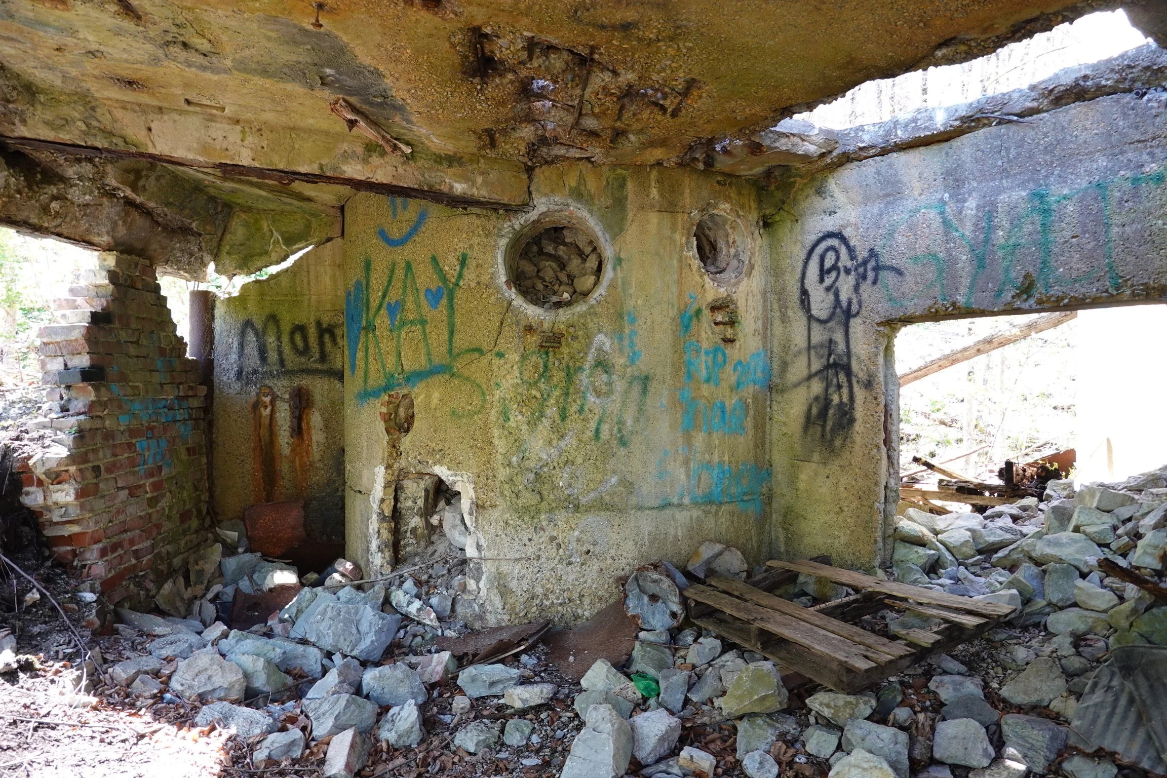 Inside the ruins of an old building with graffiti on the walls, rubble on the floor, and a large hole in the ceiling allowing light to come through.