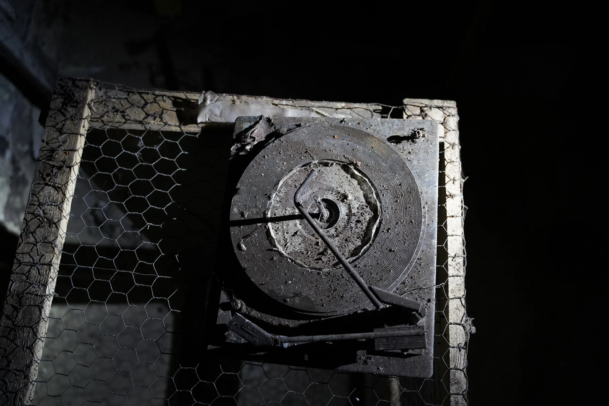 A dusty, old metal electrical timer or motor mounted on a concrete block, with wire and a hook attached, surrounded by chicken wire fencing.