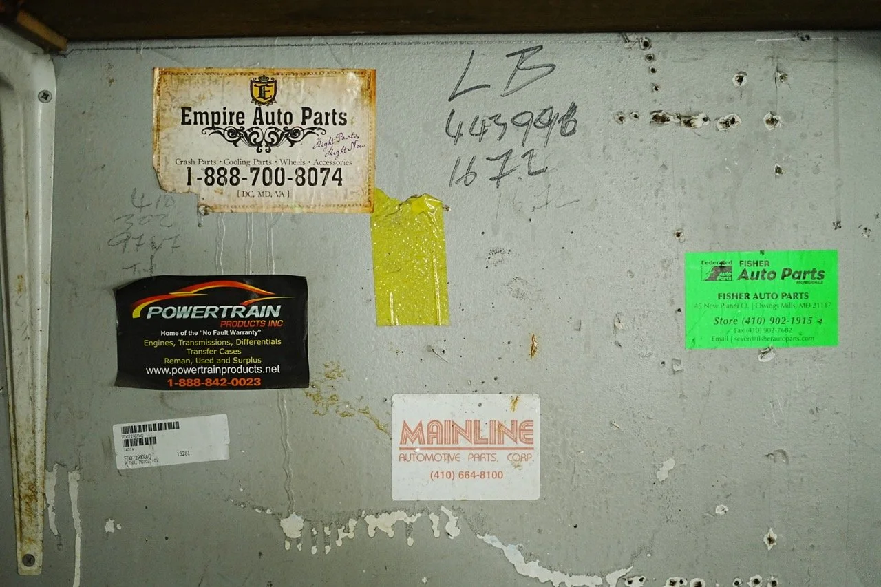 Close-up of a metal surface with various automotive parts business stickers and handwritten notes, including green, black, white, and yellow labels.