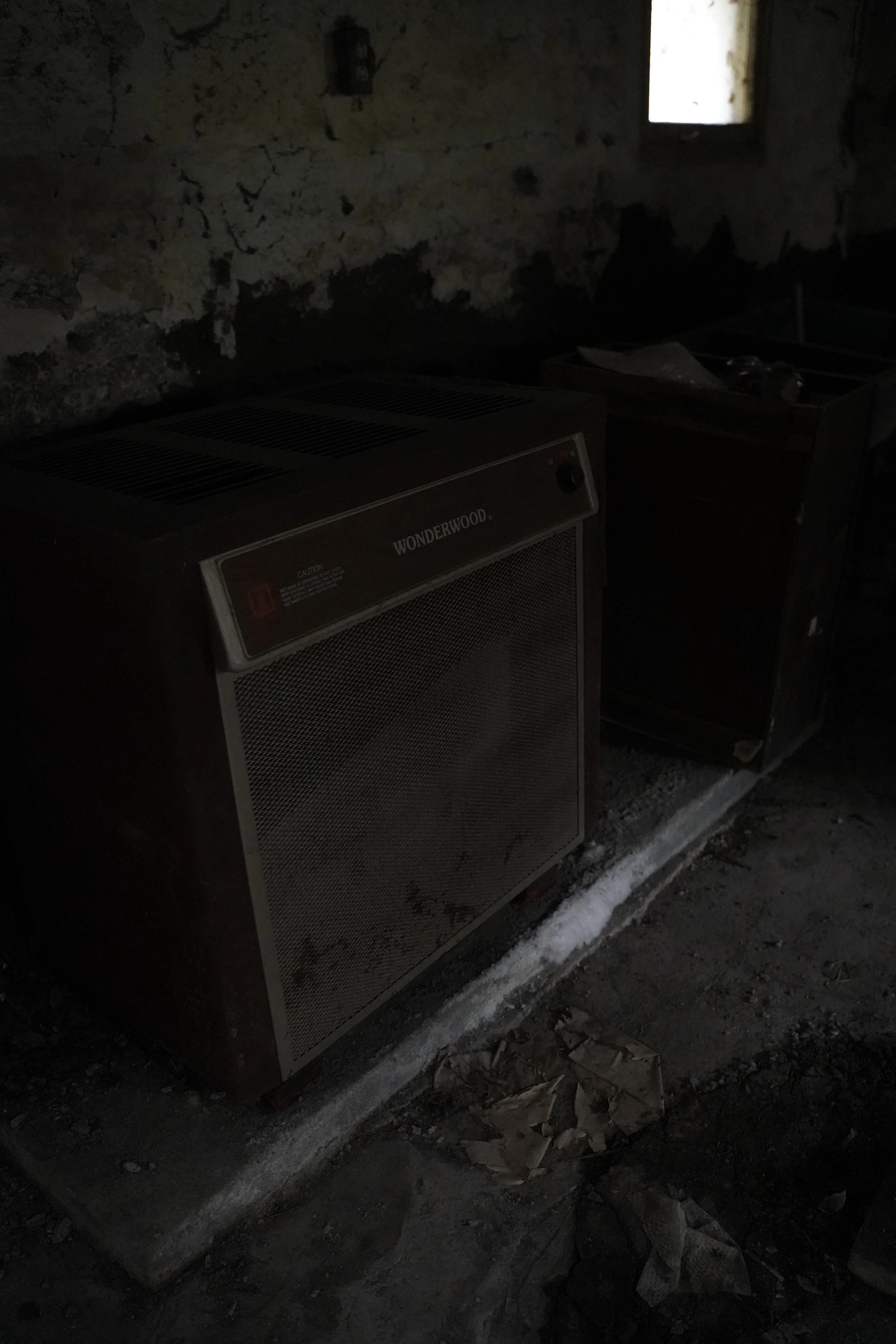 Dark indoor space with a Wonderwood heater and a small window providing minimal light, dirty floor with debris.