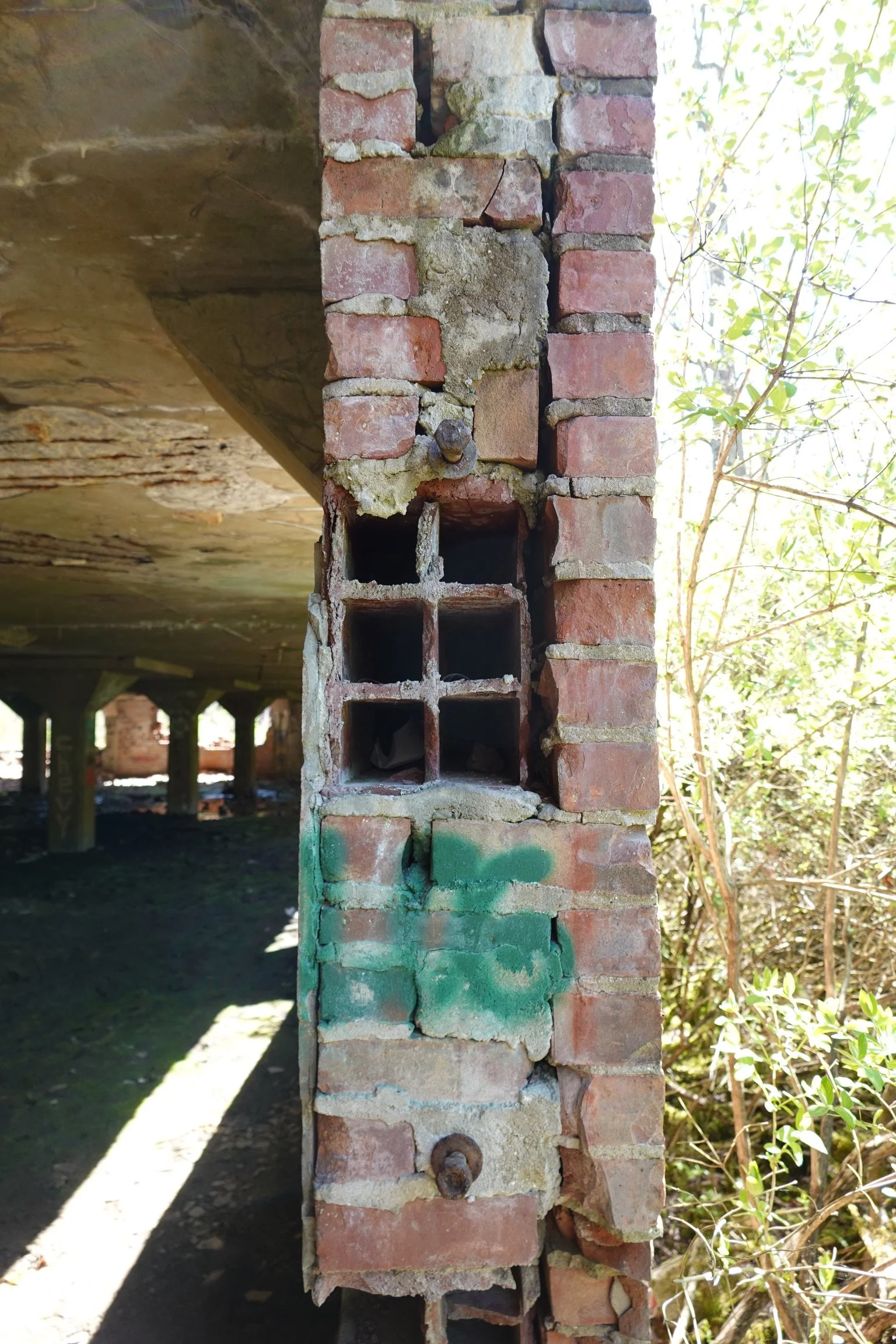 Close-up of old, weathered brick wall with exposed hollow bricks, rusted metal hooks, and greenery on the right side.