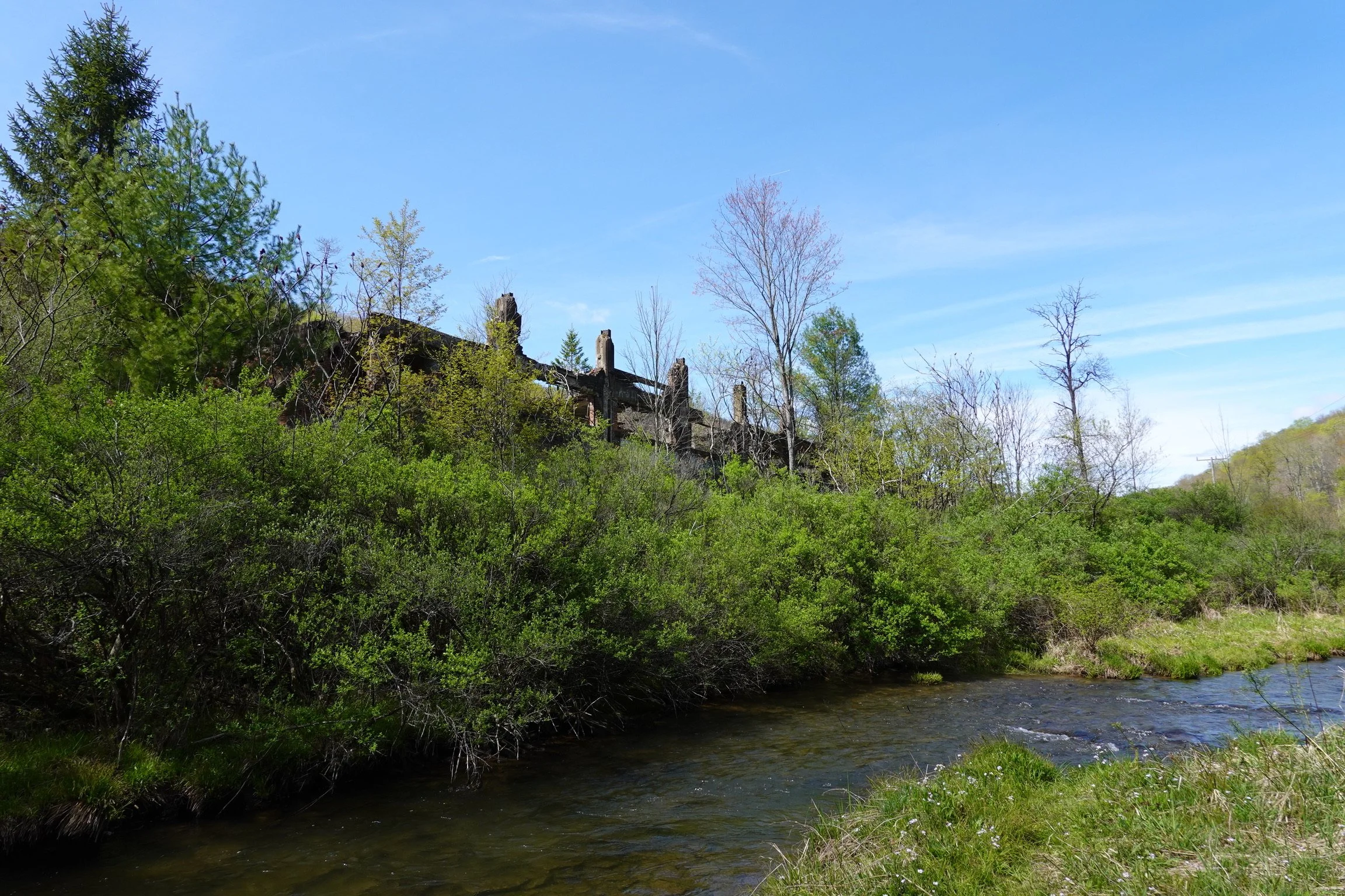 A scenic view of a river, surrounded by lush green trees and bushes, with abandoned wooden structures on a hillside in the background under a clear blue sky.