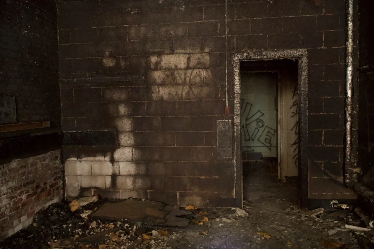 Dark, abandoned room with burned walls, debris on the floor, and an open doorway leading to another graffitied room.