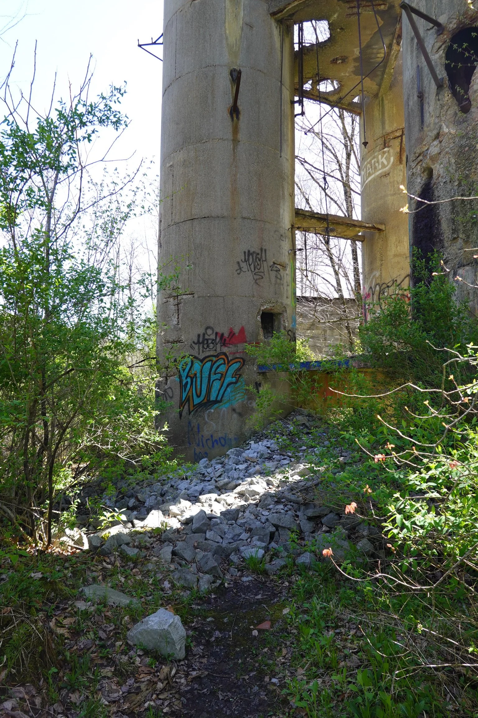 An abandoned, partially destroyed concrete structure with graffiti, overgrown with plants and surrounded by rocks and dirt.