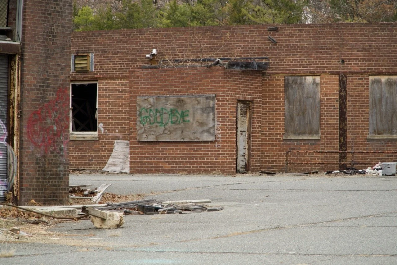 An abandoned brick building with boarded windows, graffiti, and debris on the ground.
