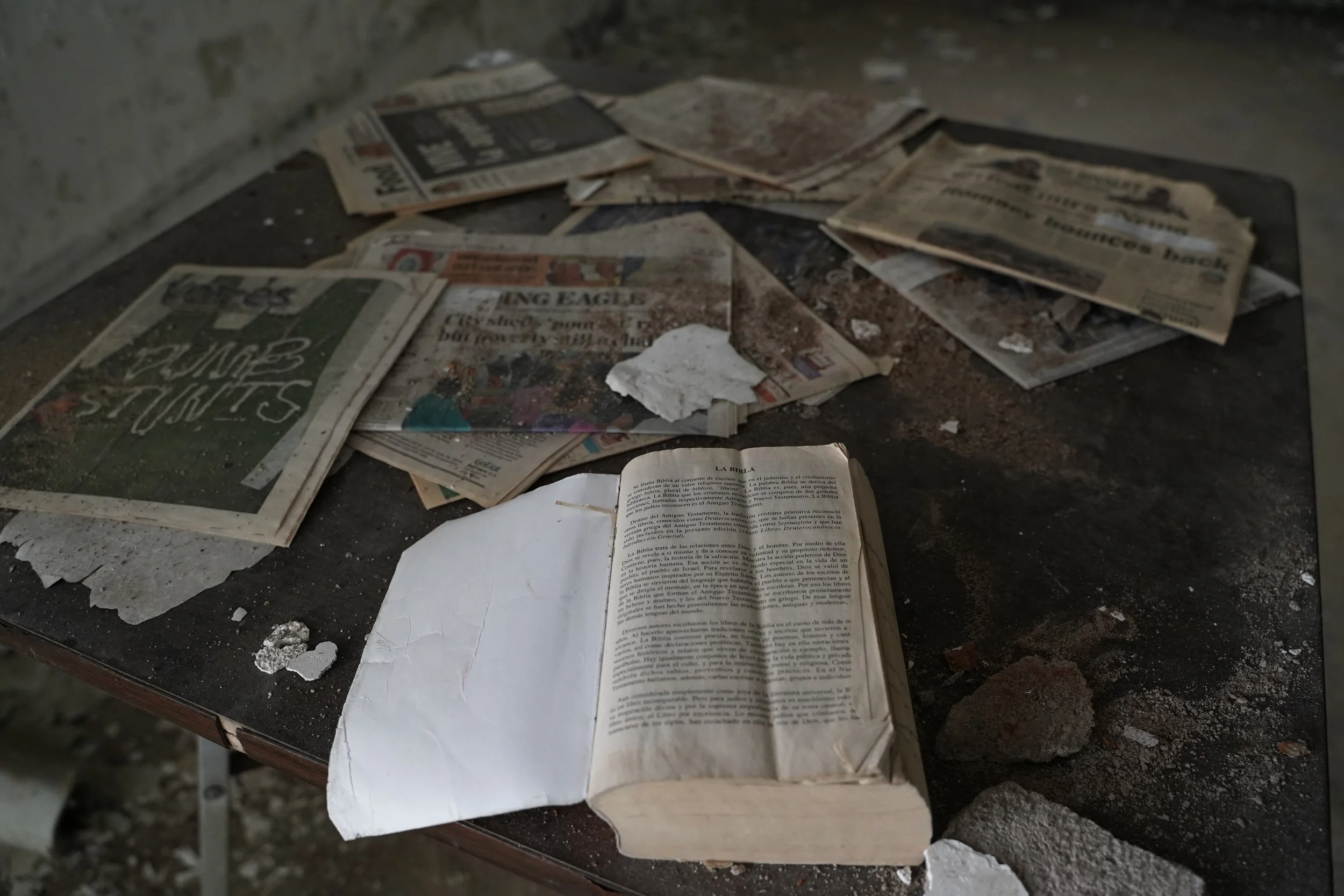 Dirty scattered newspapers and an open Bible on a dusty surface in a neglected space.