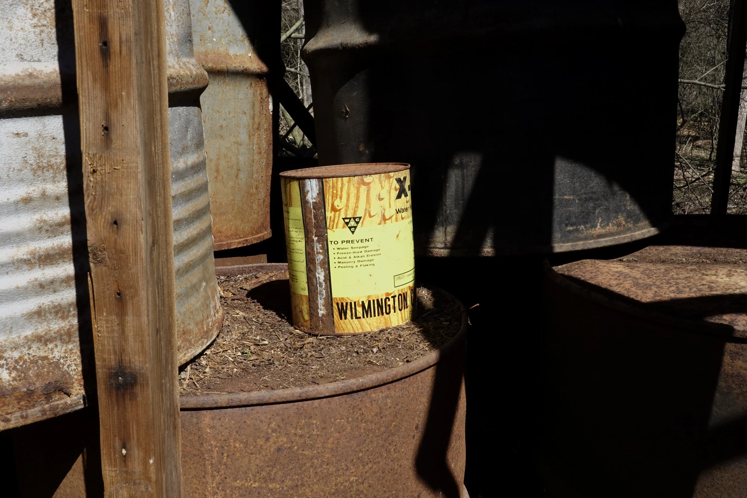 Rusty yellow metal barrel with warning label, surrounded by rusty barrels and wood posts, outdoors in a wooded area.