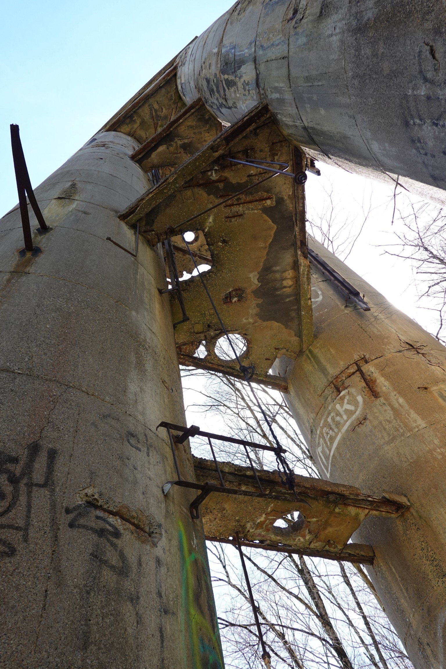 Dilapidated, abandoned industrial or factory structures with rusted metal supports and large concrete pipes, in a possibly outdoor setting with leafless trees.