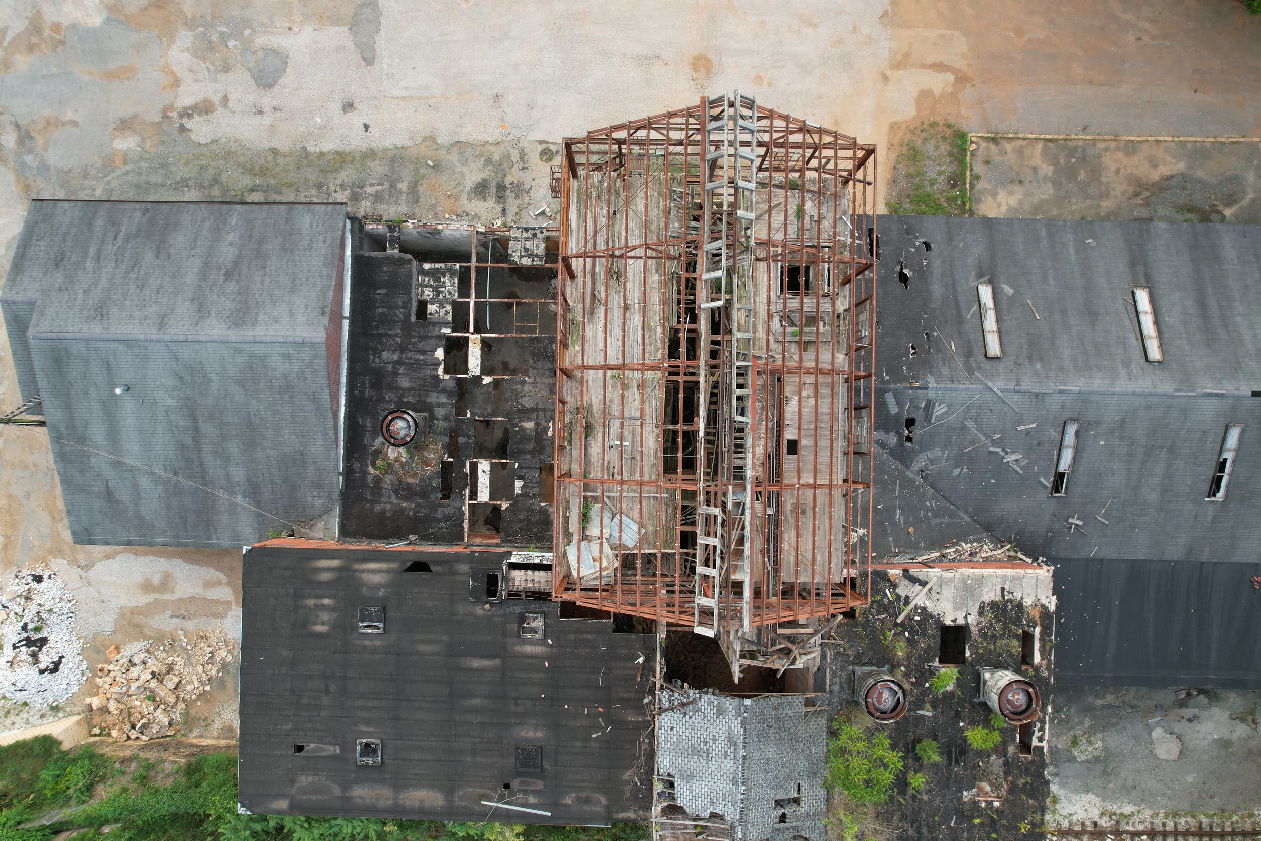 Aerial view of a building under construction with a visible steel framework, surrounded by a paved area and some greenery.
