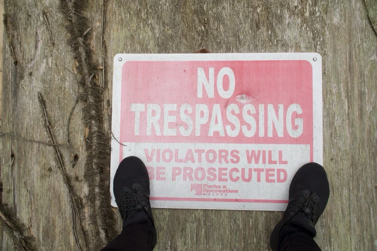 Footwear standing on a pink and white sign on a wooded surface that reads 'No Trespassing. Violators Will Be Prosecuted,' issued by Parks & Recreation.