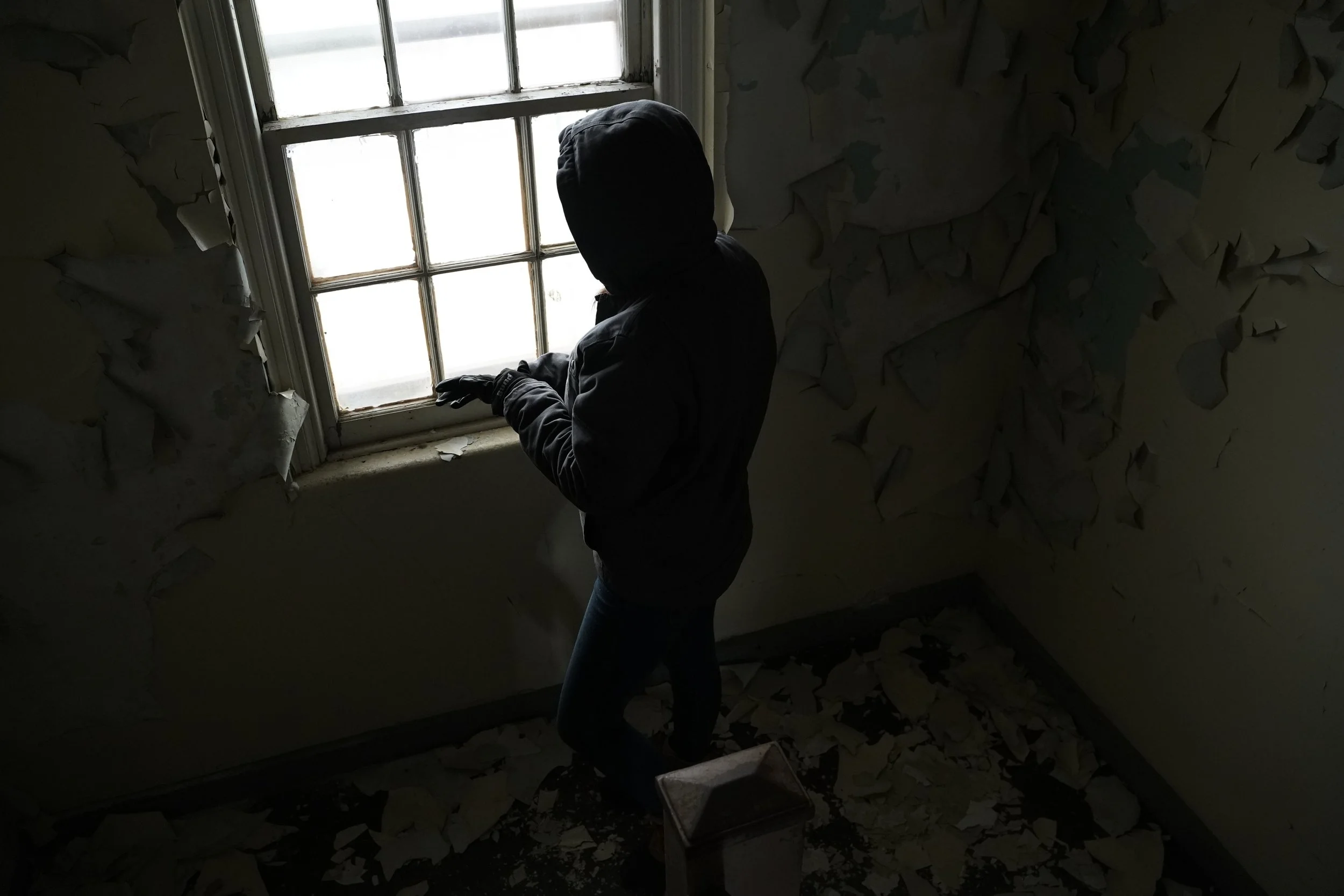 Silhouette of a person in a hoodie standing by a window in a dilapidated room with peeling paint and debris on the floor.
