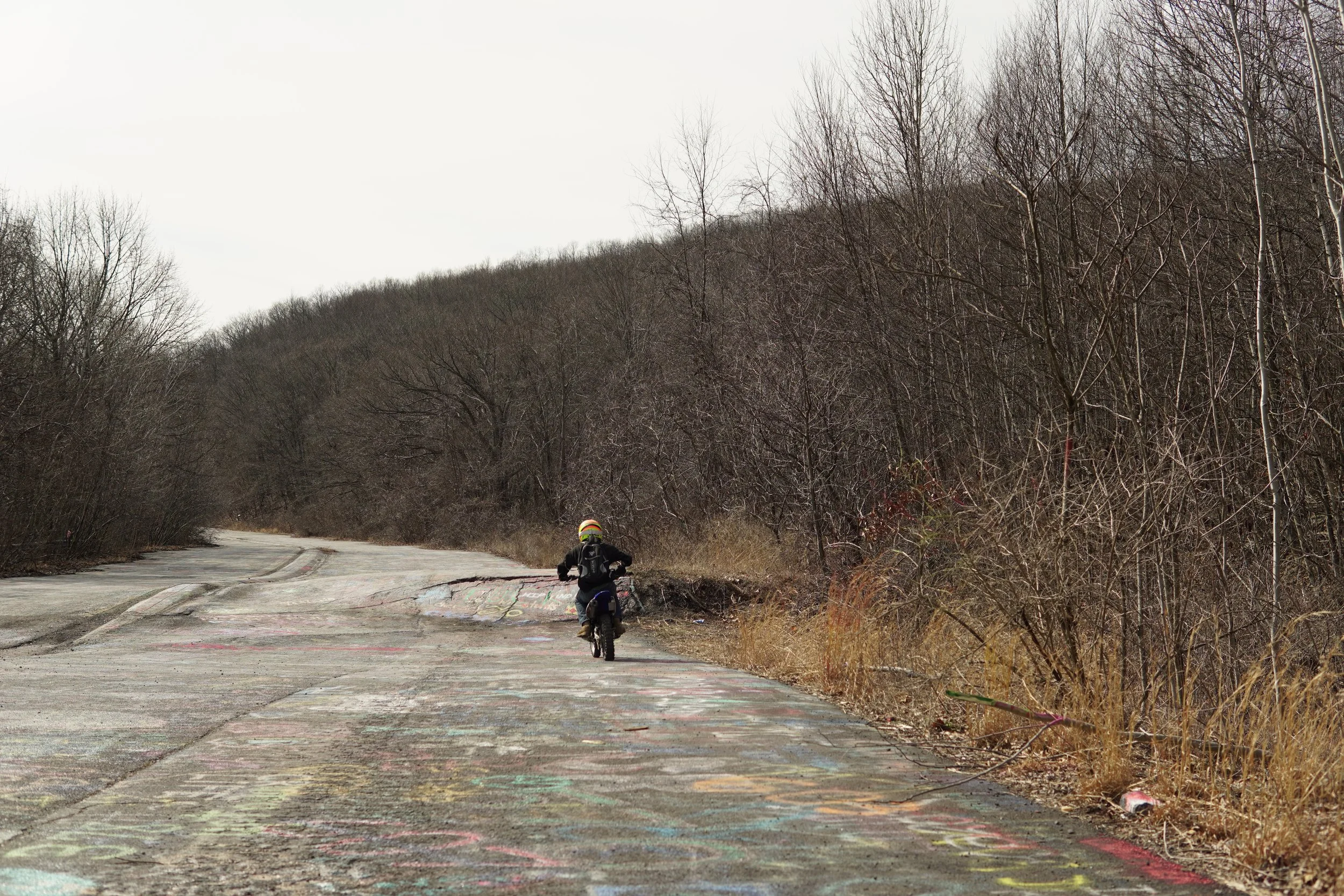 A child riding a bicycle on a worn outdoor cement path, surrounded by leafless trees and dry grass, in a rural or forested area on a cloudy day.