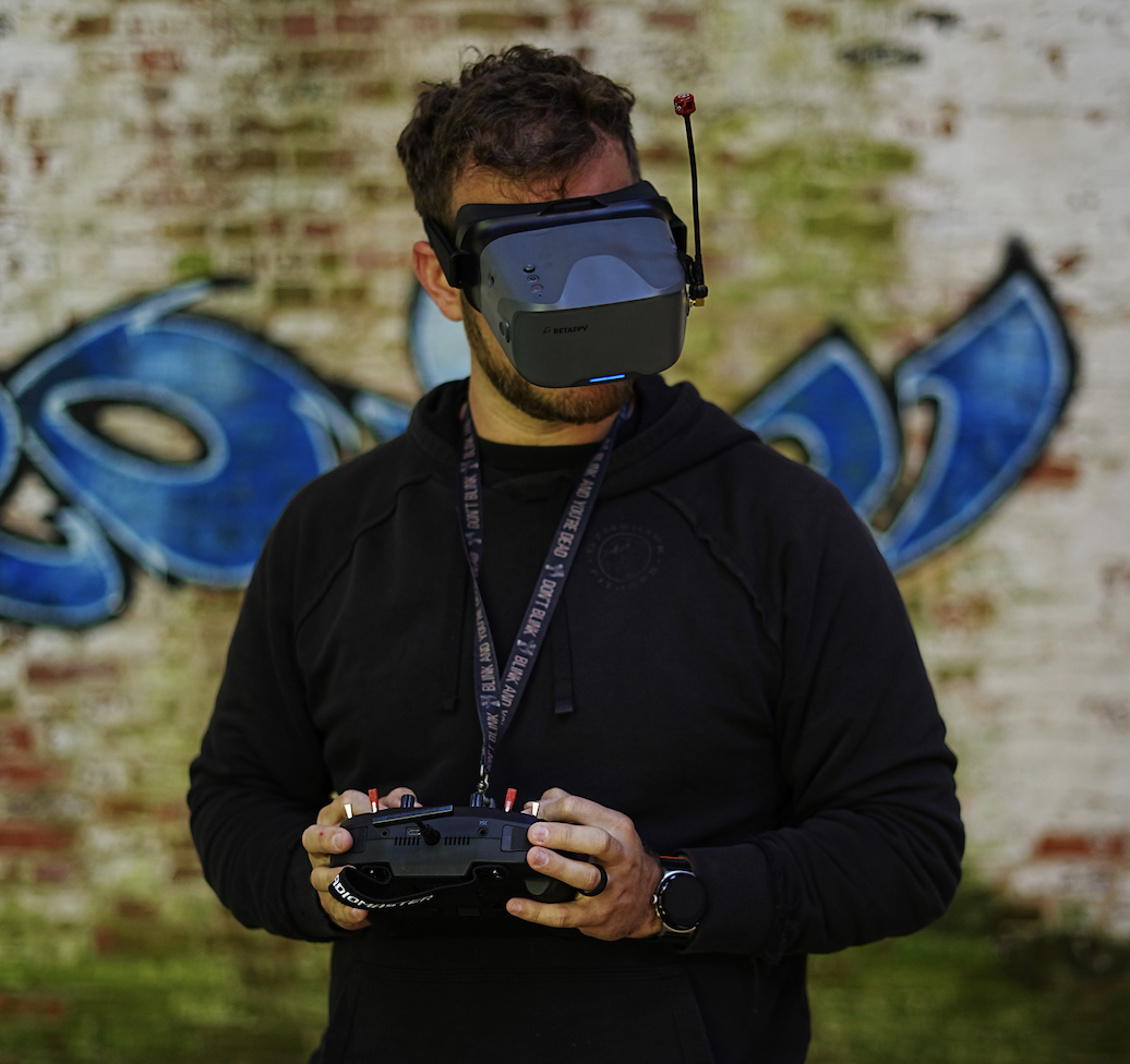 A man wearing a virtual reality headset and holding a remote control, standing in front of a brick wall with graffiti.
