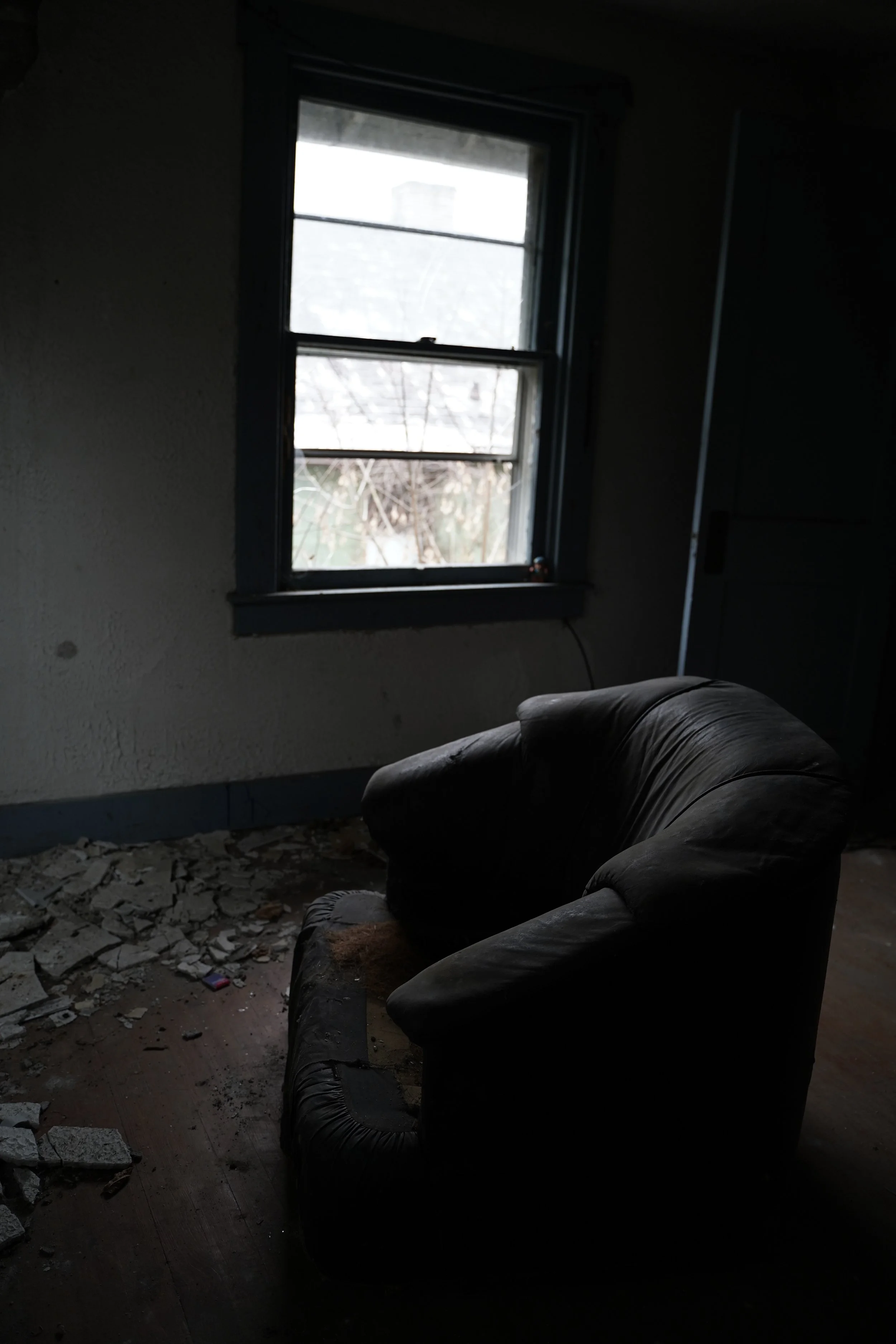 Dark living room with a black leather couch, a window showing an outdoor view, and debris scattered on the wooden floor.