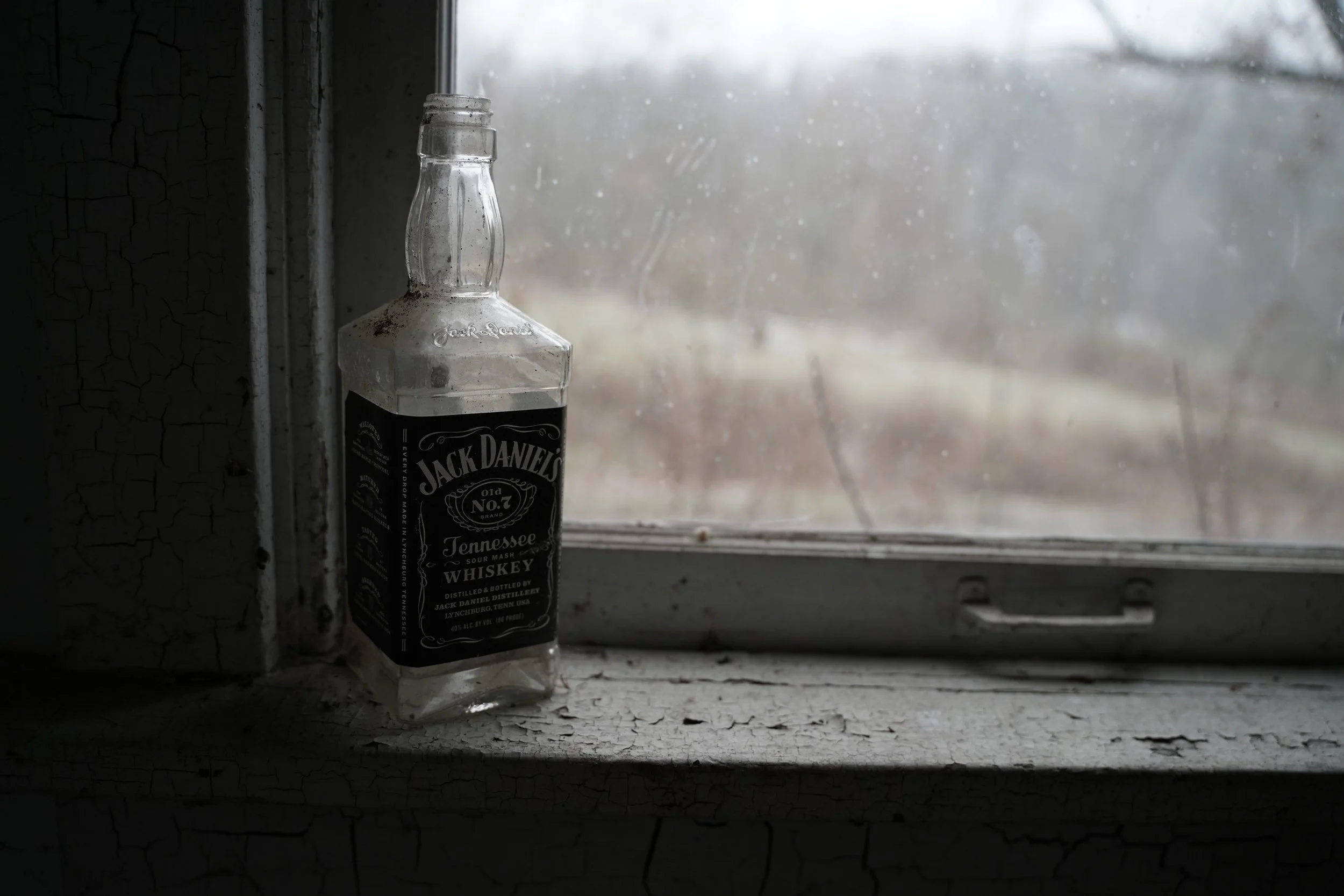 A nearly empty Jack Daniel's Tennessee Whiskey bottle on a windowsill, with a foggy outdoor scene visible through a dirty window.