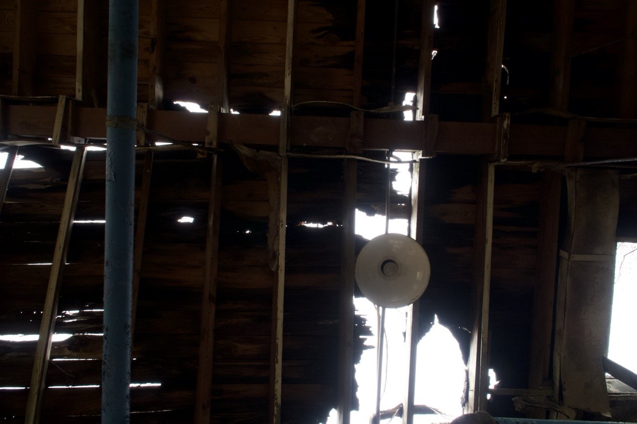 A broken wooden ceiling with gaps and torn planks, sunlight shining through the gaps, a blue pole in the foreground, and a circular light fixture hanging from the ceiling.