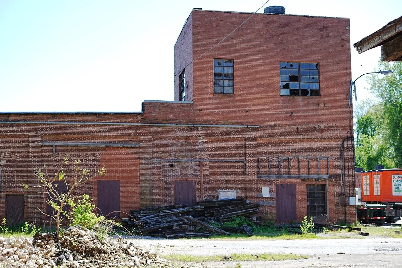 An old, abandoned brick building with broken windows and boarded-up doors, surrounded by gravel and some sparse greenery.