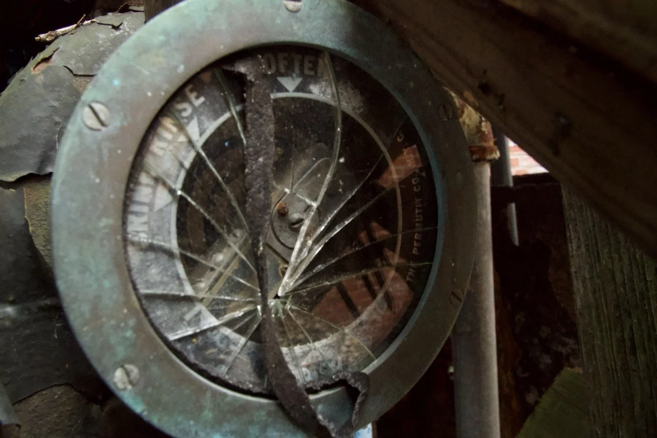 Close-up of a broken, shattered circular gauge or instrument panel with cracked glass, rusted metal frame, and dusty, worn surface.