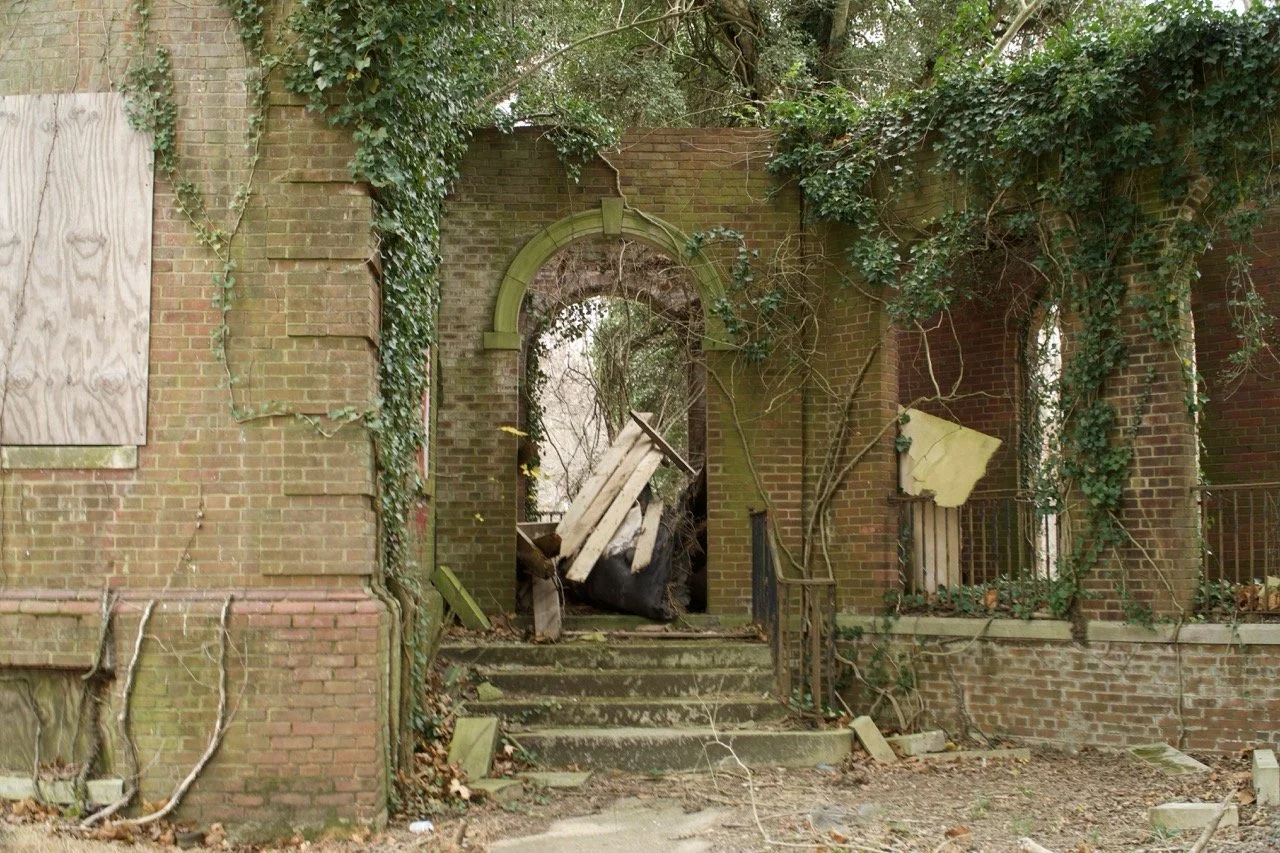 An abandoned brick building with overgrown vines, a boarded-up window on the left, a set of steps leading to an arched doorway filled with debris, and broken or missing panels on the walls.
