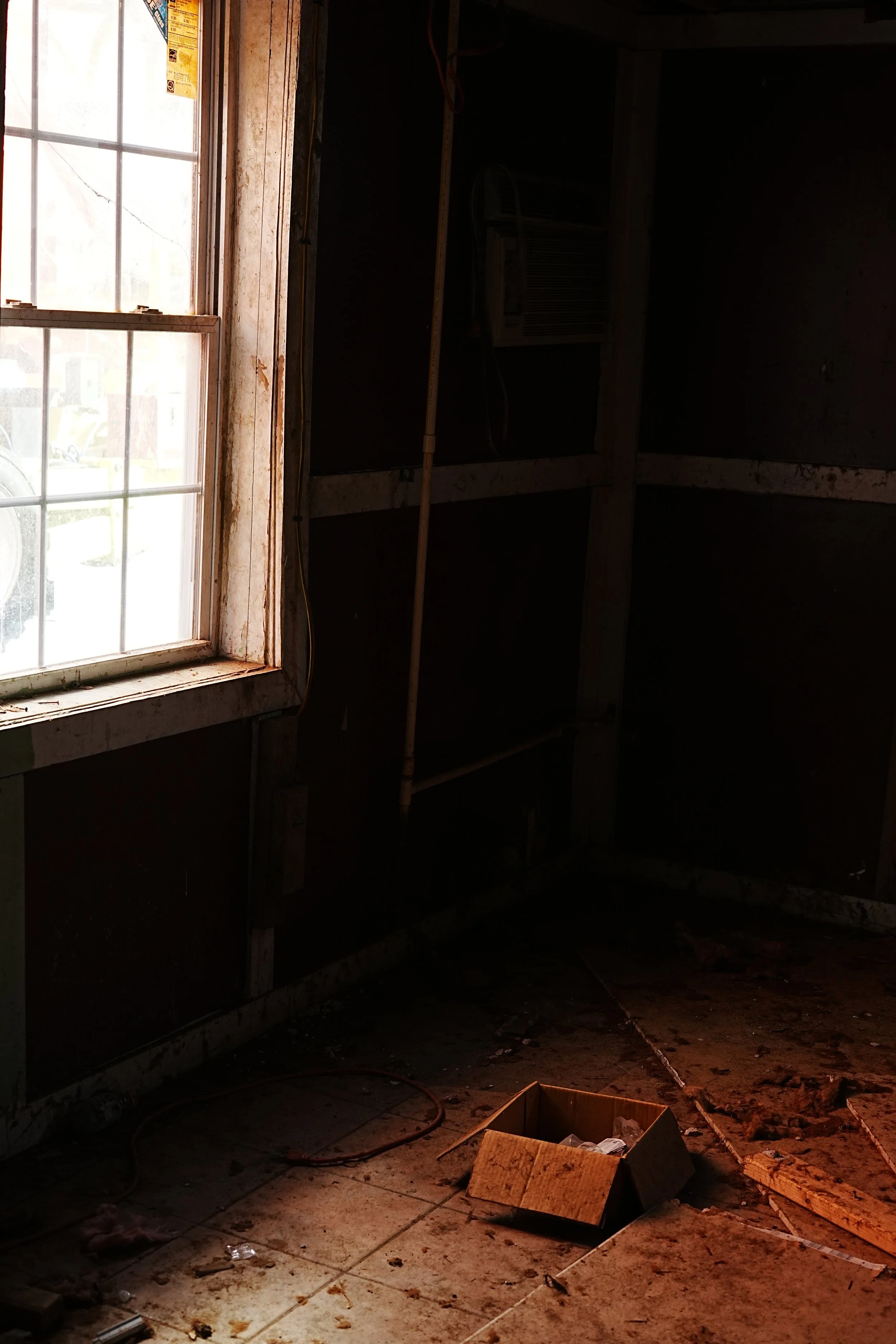 A dimly lit construction or renovation site with a window on the left, scattered debris, a cardboard box on the floor, and construction materials and framing visible.