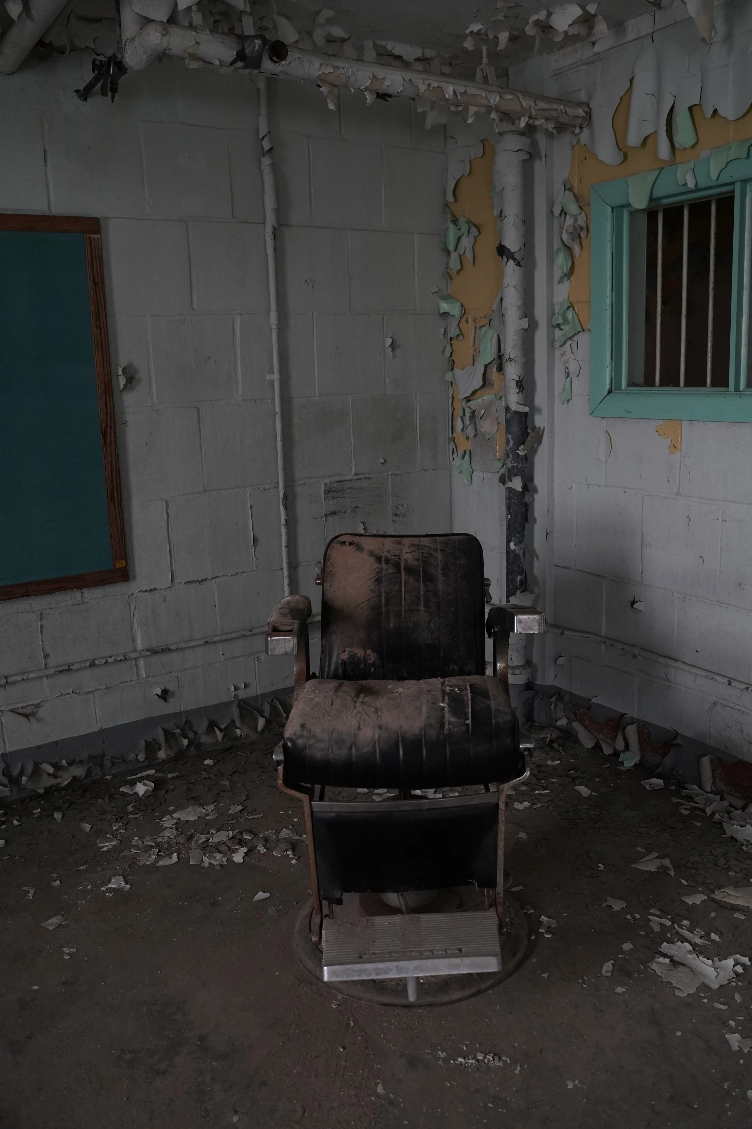An abandoned, dusty barber chair in a neglected room with peeling paint, debris on the floor, a barred window, and a concrete block wall.