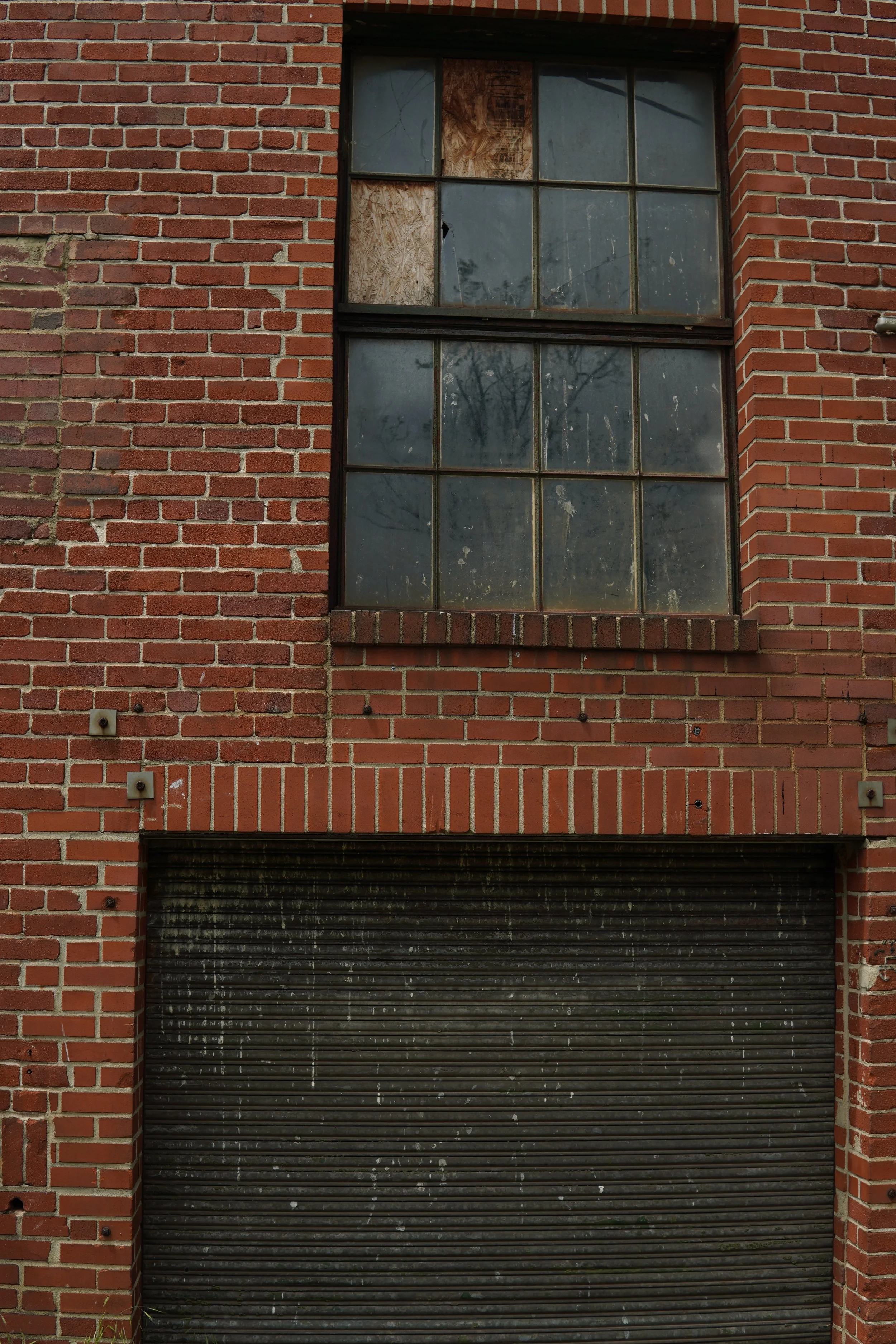 The image shows a brick building with a large, grid-style window that is partly boarded up and dirty, and a black, closed rolling garage door beneath the window.