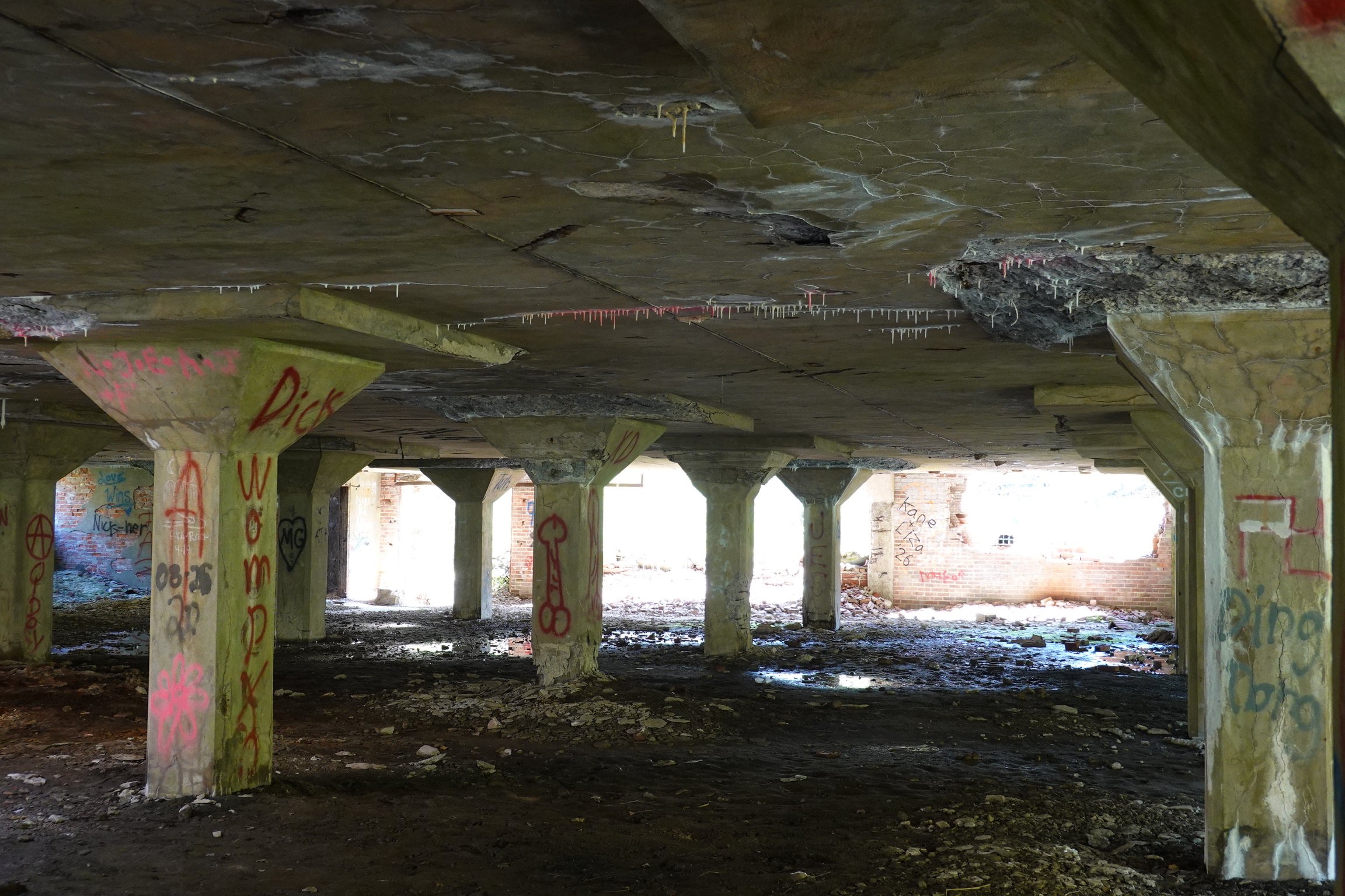 An abandoned building with graffiti-covered concrete pillars and walls, a dirt floor, and a ceiling with water stains and cobwebs. Bright light appears to be coming from the openings in the building.