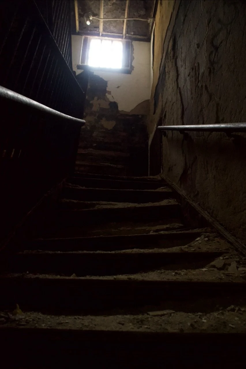 Dimly lit, dusty staircase inside an old building with peeling walls and a window at the top allowing bright light to shine through.