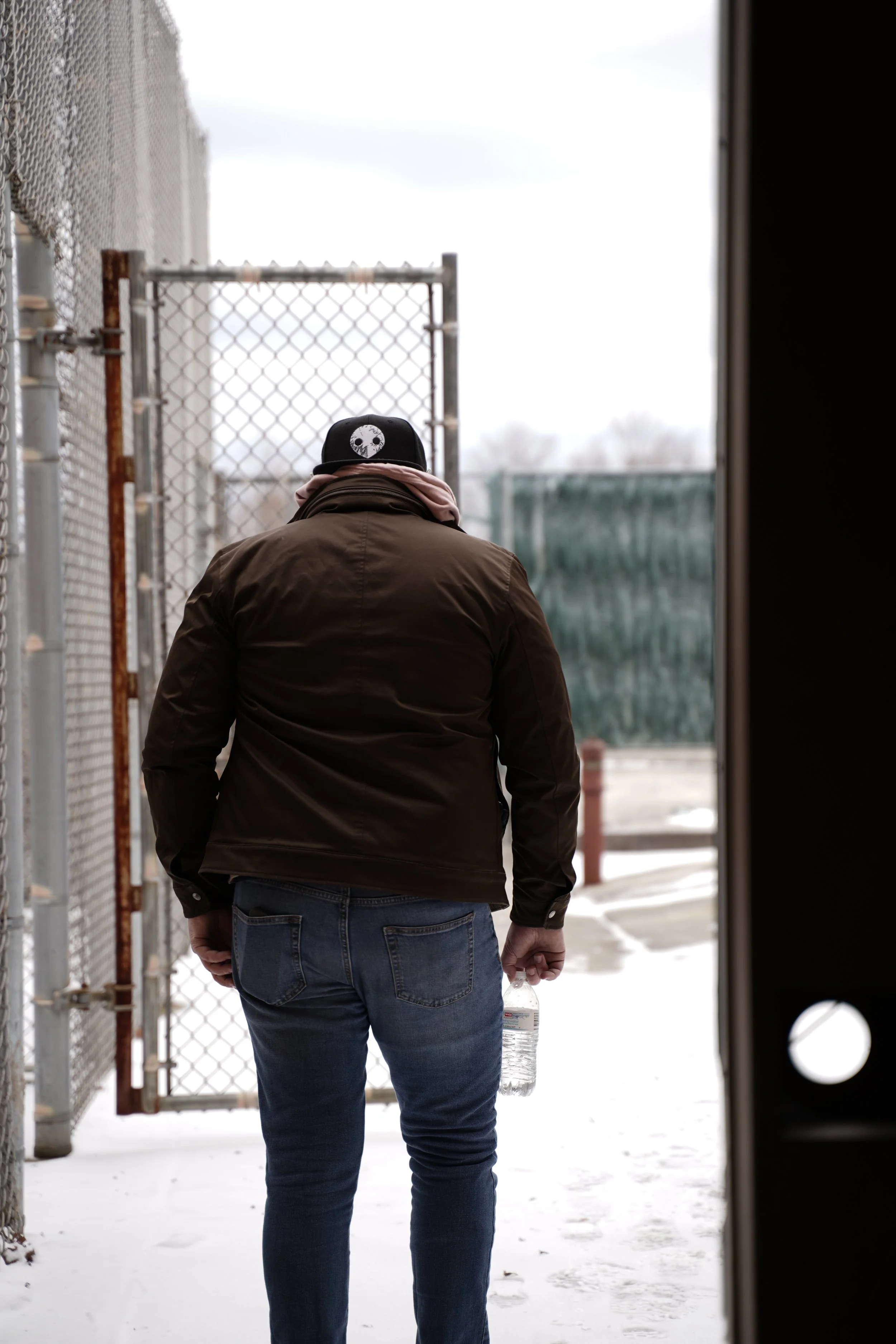 A person dressed in a brown jacket and blue jeans walking through a snowy alleyway, holding a water bottle, with a chain-link fence on the left and a black hat with a skull design on their head.