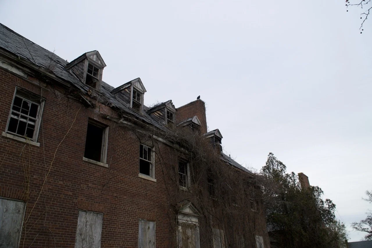 An old brick building with boarded-up windows and missing roof shingles, some windows are open, and there are trees surrounding the building, under a cloudy sky.