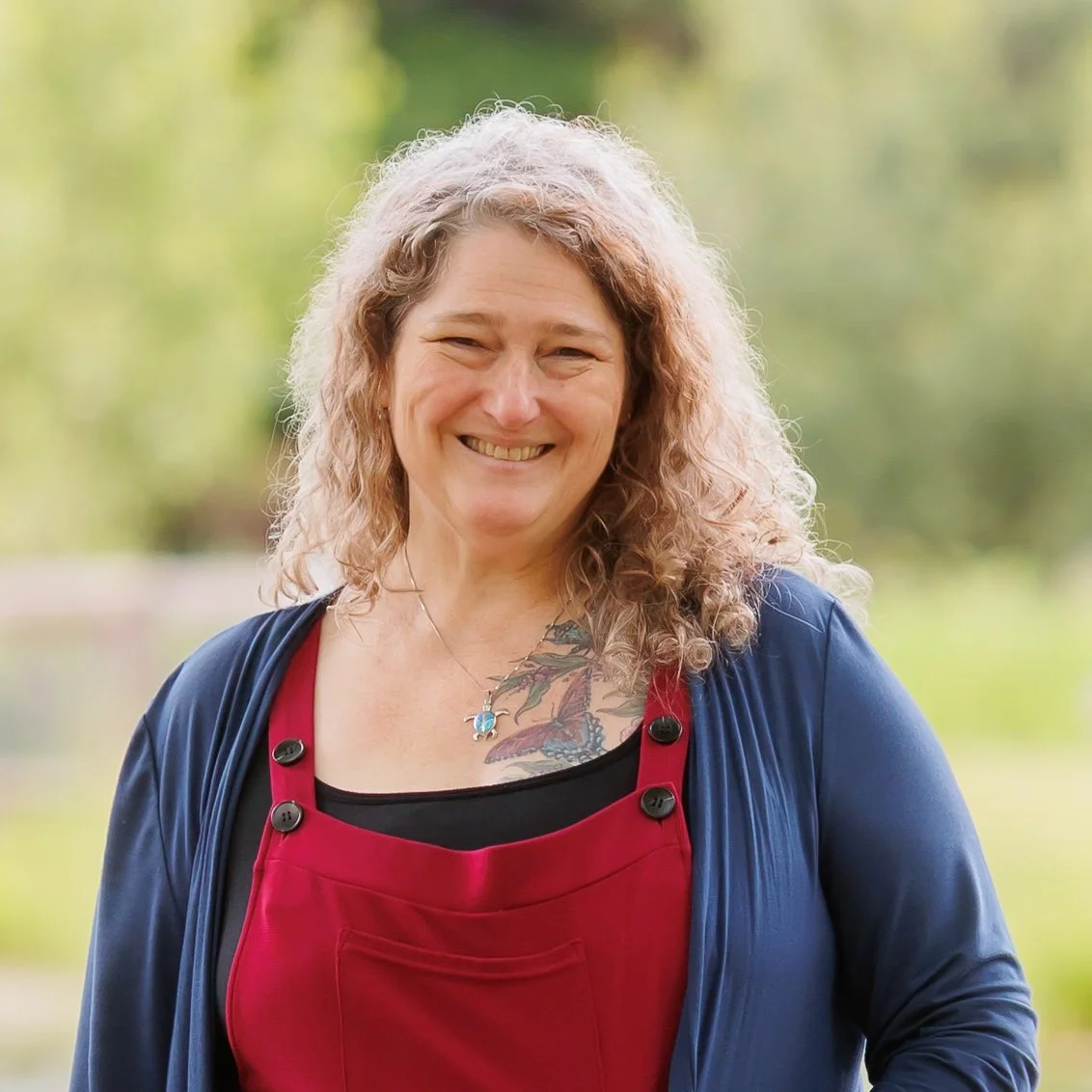 A smiling woman with curly blonde hair outdoors, wearing a red apron over a black shirt and a blue cardigan, with a tattoo on her chest and a necklace with a turquoise pendant.