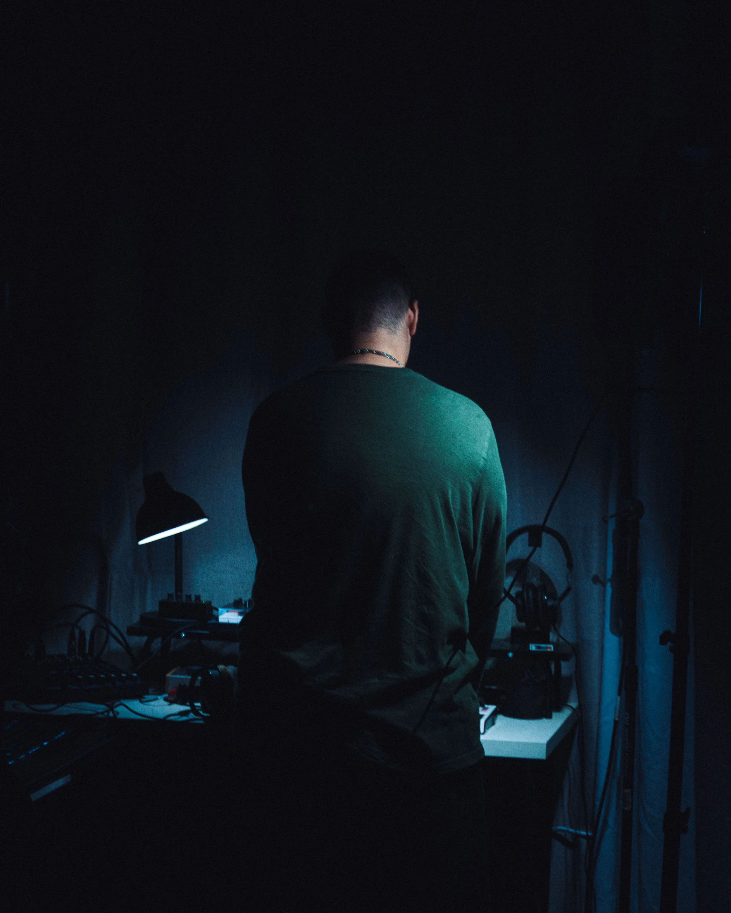 Persona con cabello corto desde atrás, usando camiseta verde, en un cuarto oscuro, parece que está mezclando música con equipos de DJ, rodeado de cables y dispositivos electrónicos.