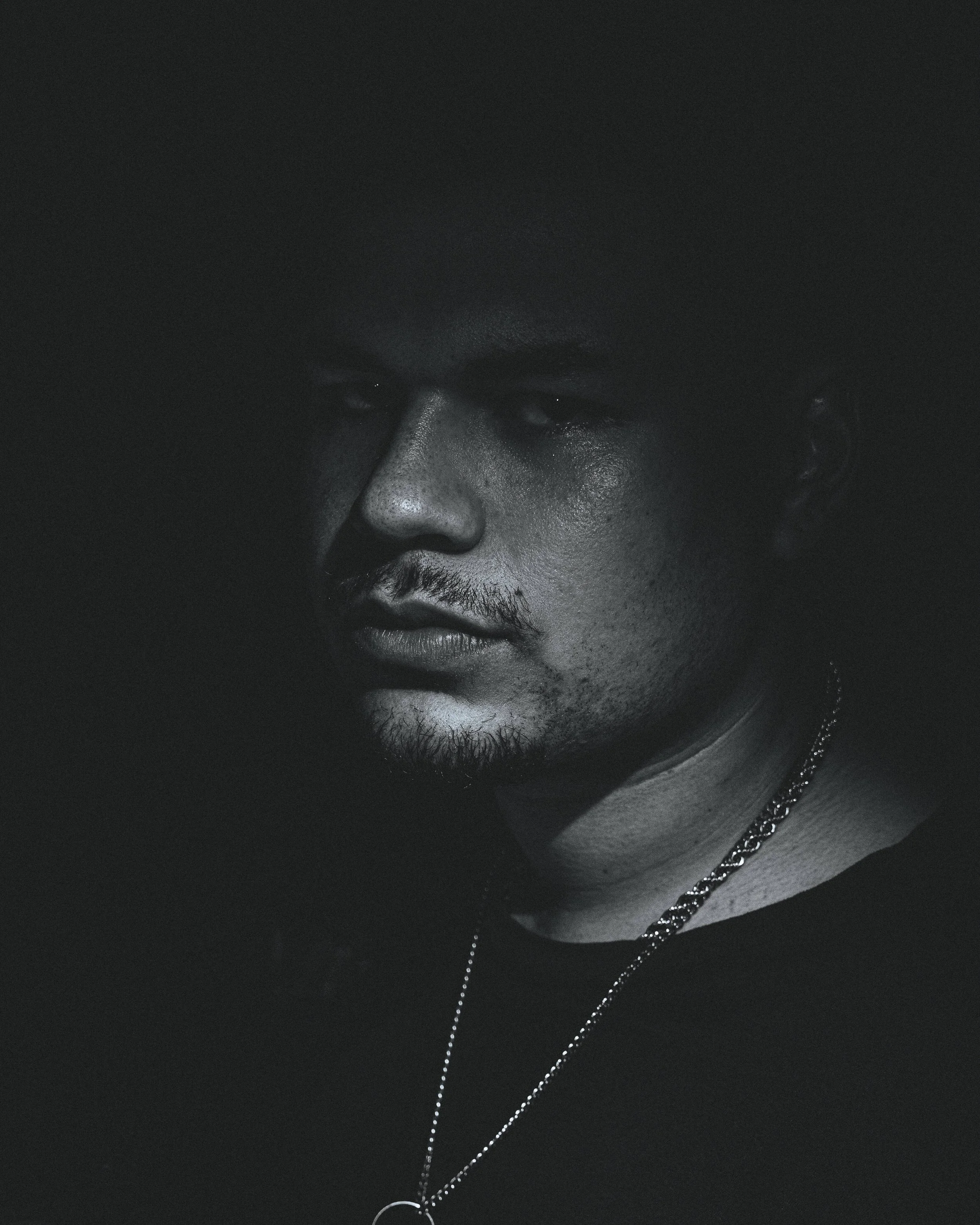 Primer plano de un rostro masculino en tonos oscuros, con expresión seria. La imagen es en blanco y negro, con poca iluminación resaltando las características faciales y cadenas de plata en el cuello.