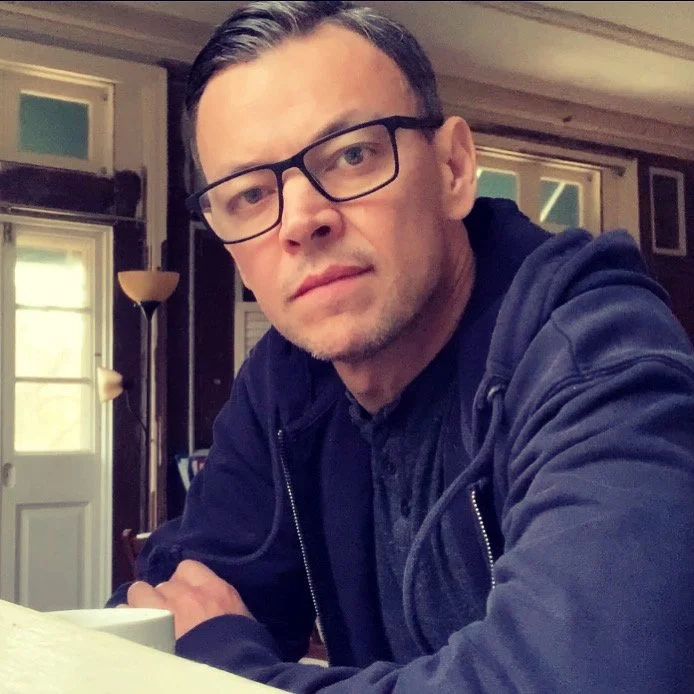 A man with glasses and dark hair, wearing a dark jacket, sitting indoors, looking directly at the camera.