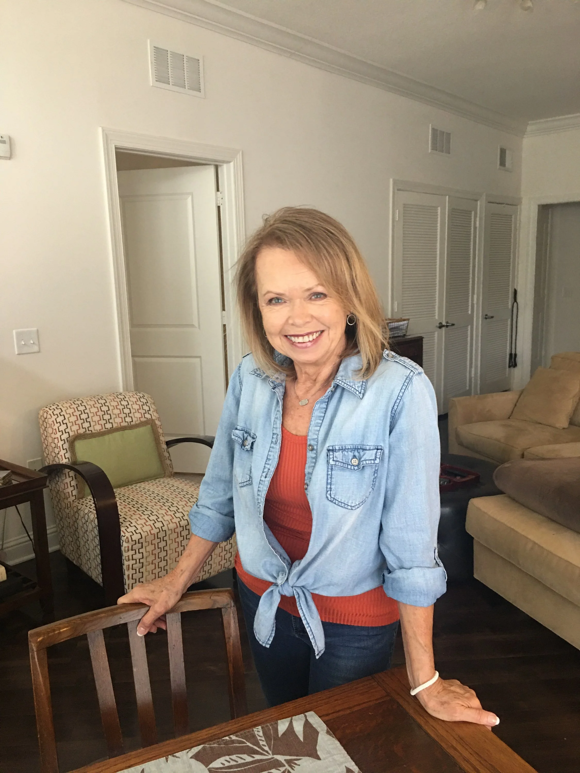 A smiling woman with shoulder-length blonde hair, wearing a denim shirt over a red top, stands in a living room with her hands resting on a wooden dining table. The room has beige walls, a patterned armchair, a beige sofa, and closed white double doors.