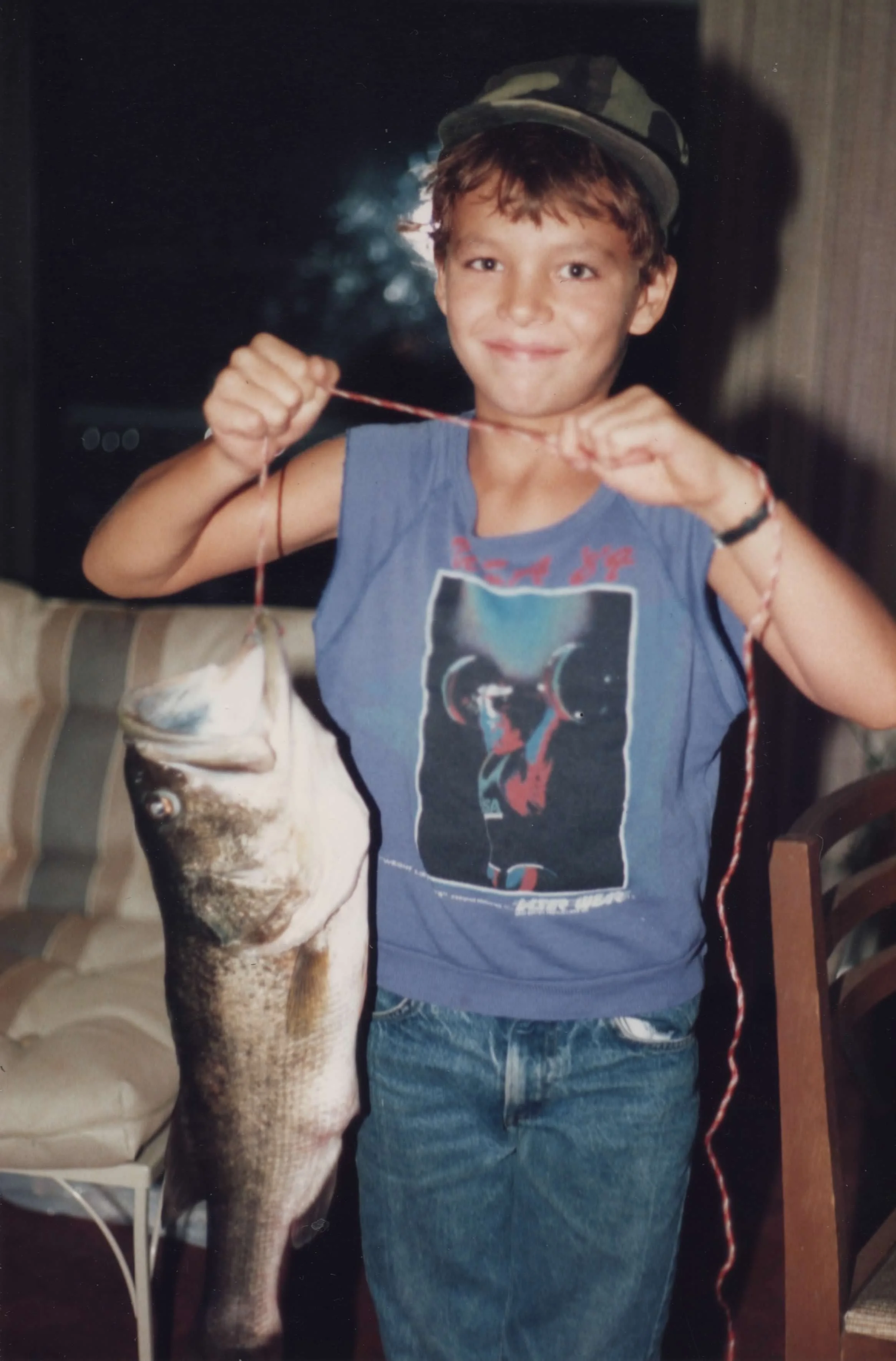 A young boy in a camouflage cap and blue sleeveless shirt holding a large fish vertically by its mouth in an indoor setting.