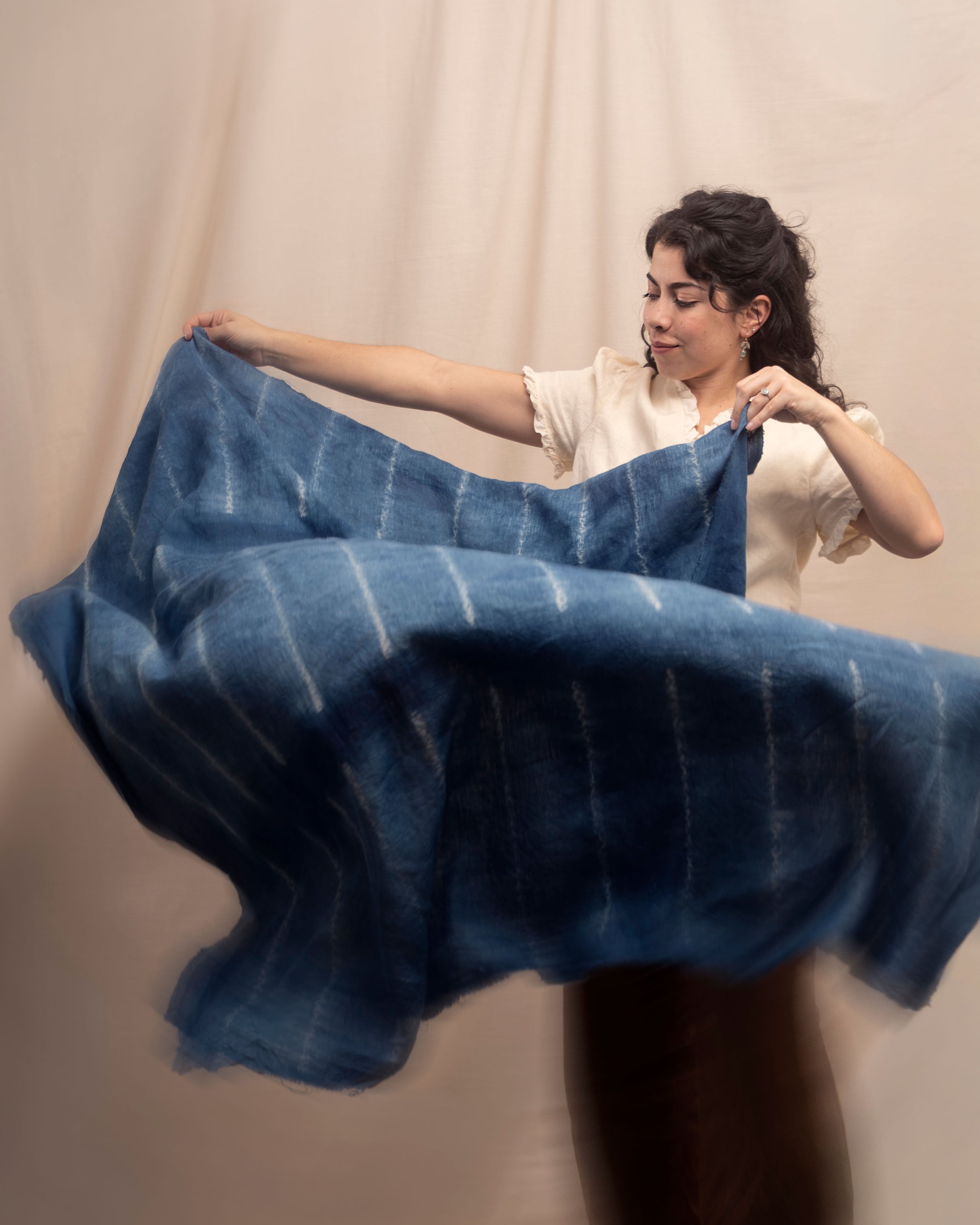 A woman with dark, curly hair and earrings holding a piece of blue fabric with white windowpane pattern, standing against a light-colored curtain background.